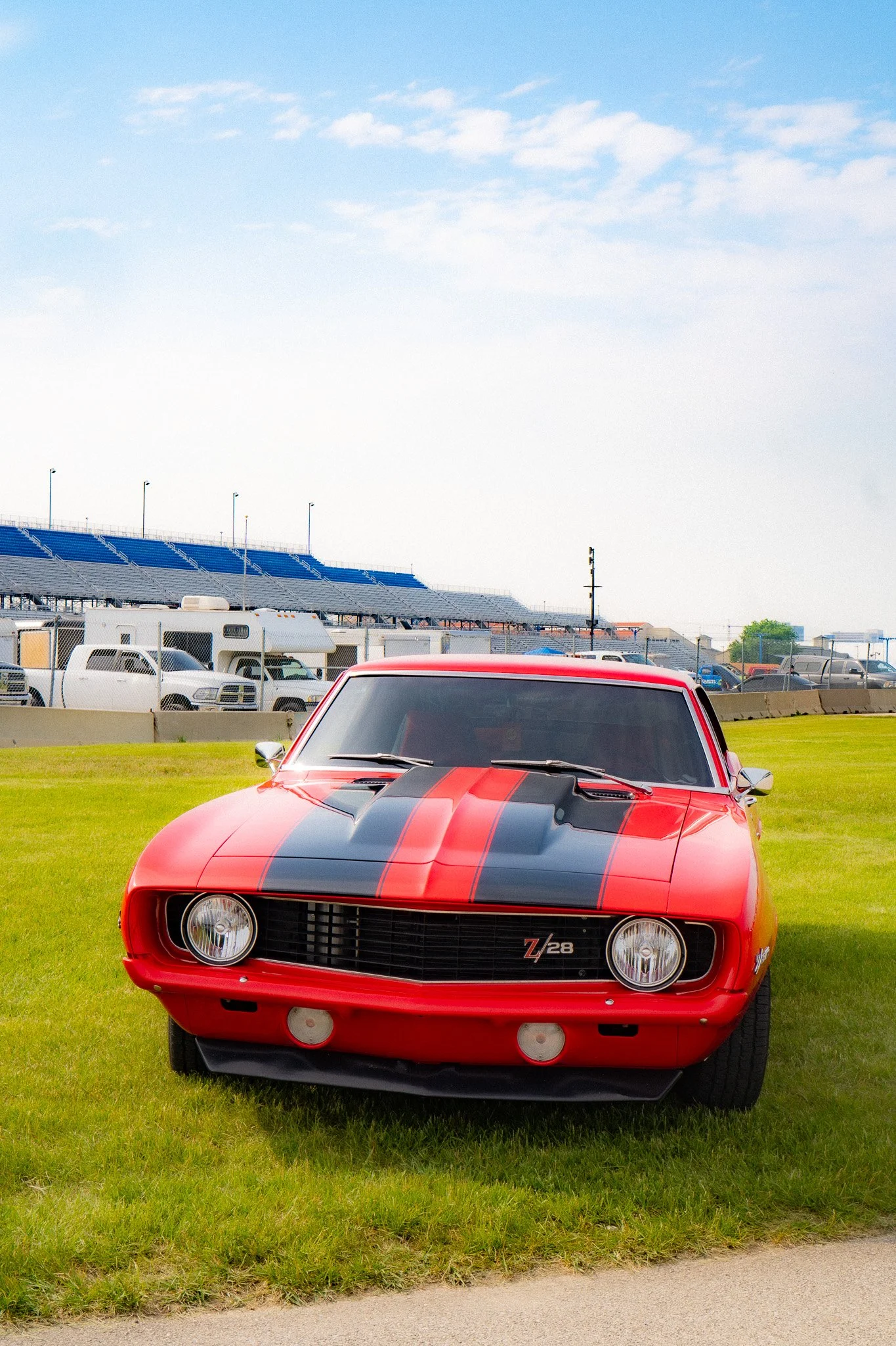 Front view of a red Chevrolet Z28 Camaro with black racing stripes, parked on grass at a race track, with bleachers, vehicles, and a blue sky in the background.