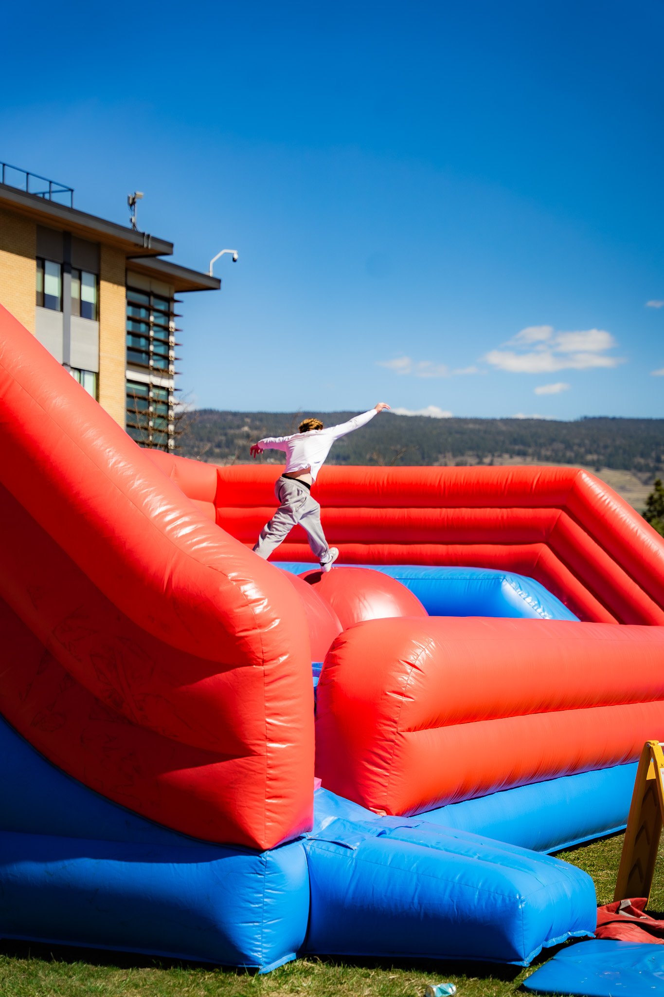 Person in white shirt and gray pants jumping on a red and blue inflatable bounce house outdoors on a sunny day with a building and mountains in the background.
