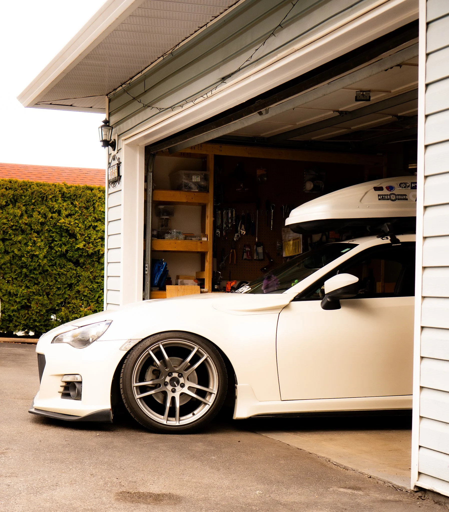 White sports car with roof cargo box partially parked in a garage, tools and shelves visible inside, with greenery outside.