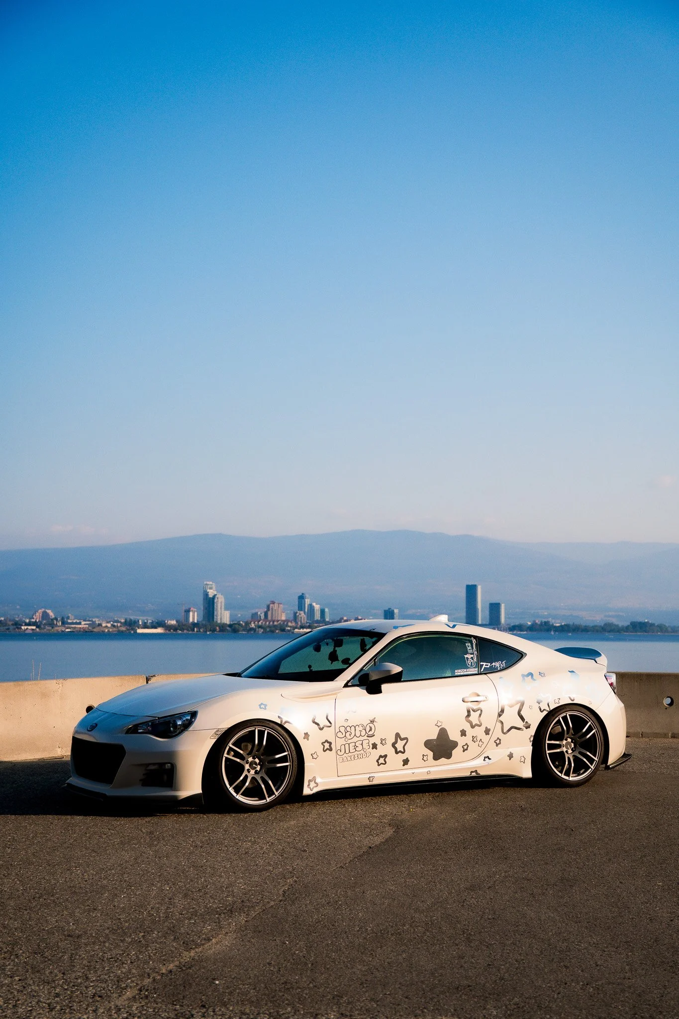 White sports car with black stars and doodles parked along a waterfront with a city skyline and mountains in the background
