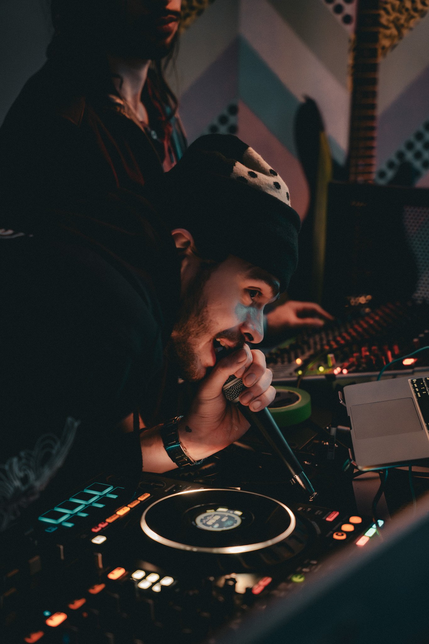 A DJ with a beard and wearing a beanie is operating a DJ mixer with many colorful lights, in a dimly lit environment, likely at a nightclub or event.
