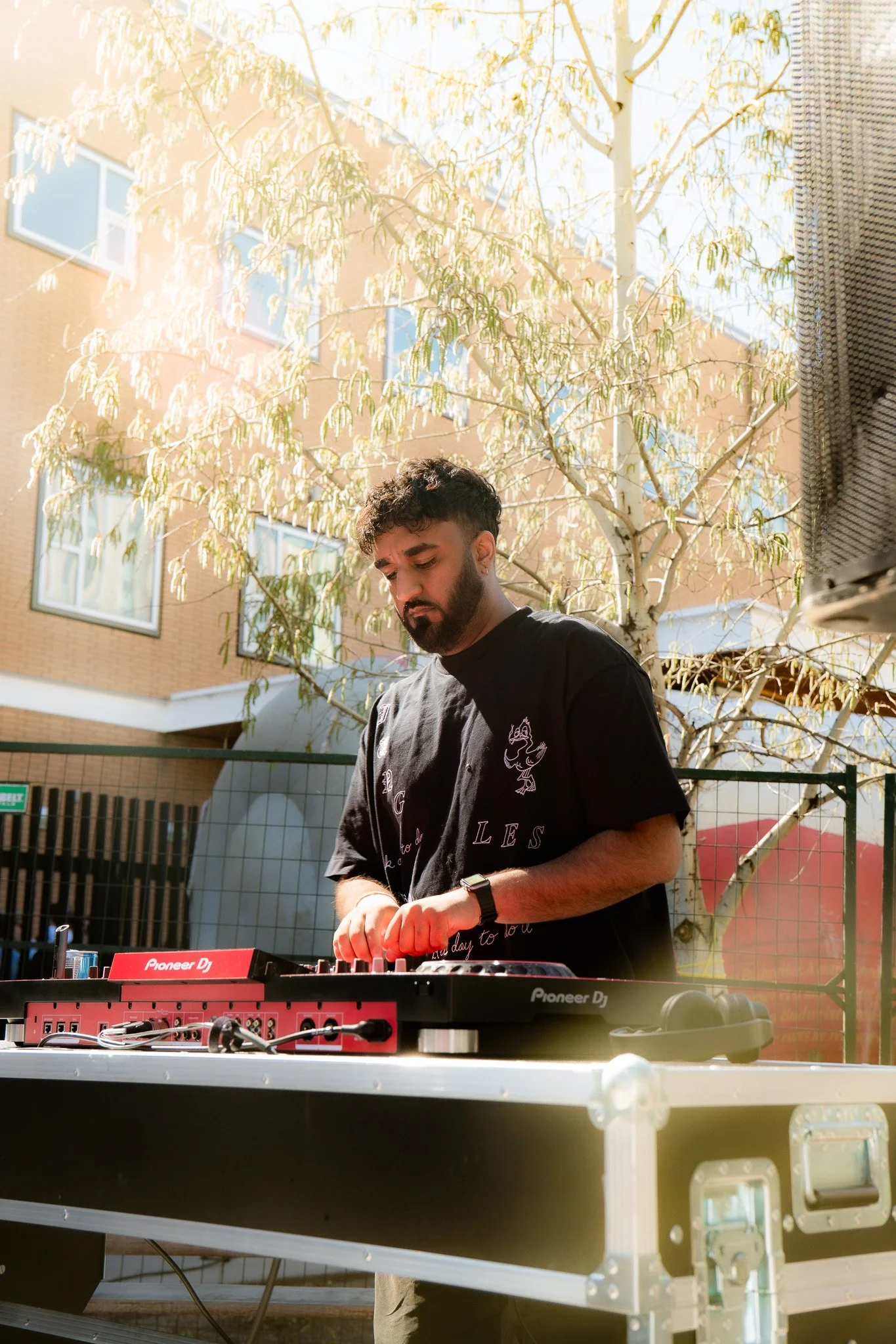 A young man with dark curly hair and a beard is DJing outdoors, wearing a black T-shirt with white text and a wristwatch on his left hand. He is using a Pioneer DJ controller, and in the background, there are buildings with windows and a leafy tree. The scene is sunlit.