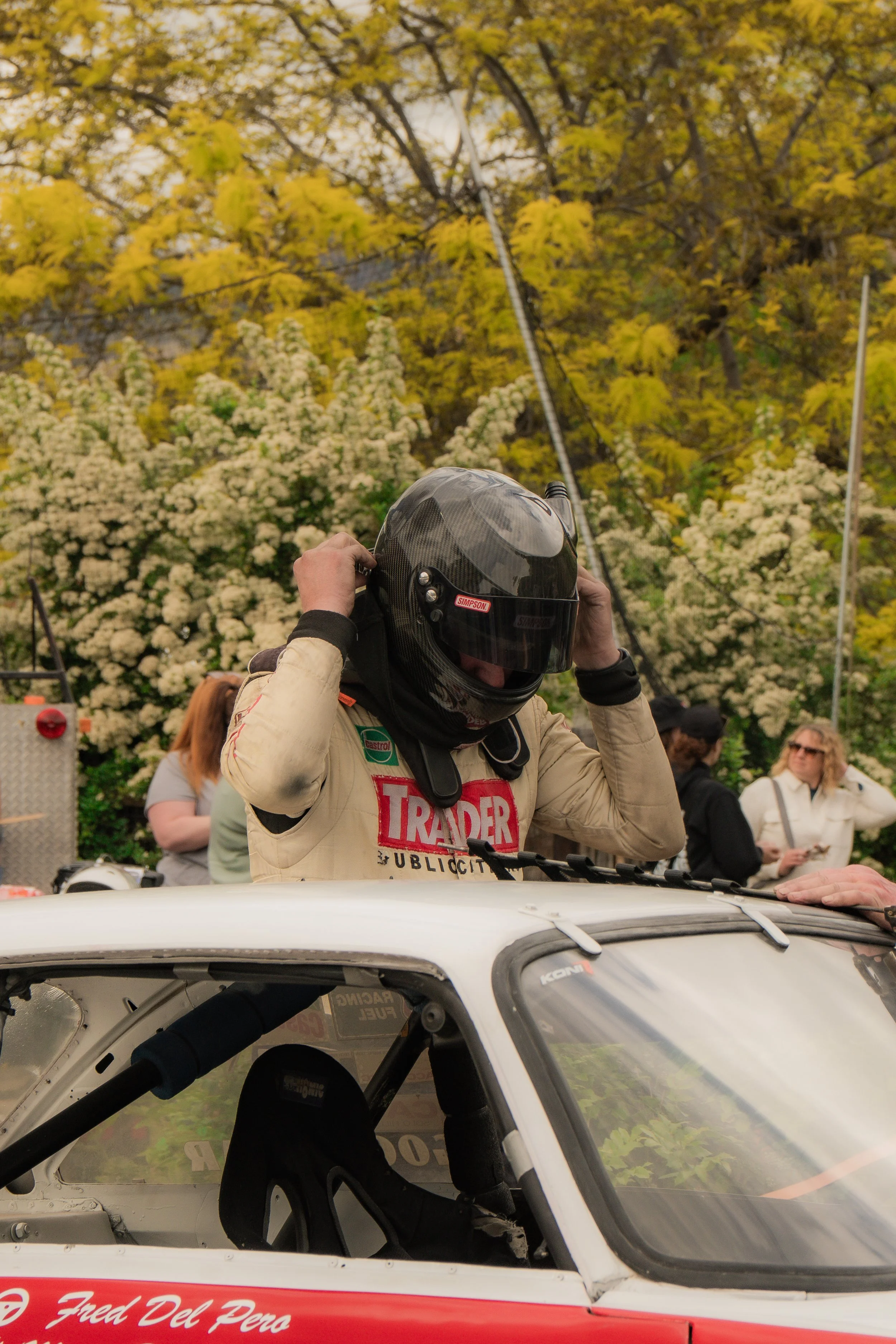 A race car driver wearing a helmet and racing suit adjusting his helmet, standing outside a white race car with a red stripe and the name 'Fred Del Peco' on the side. In the background, there are spectators and trees with yellow-green leaves.