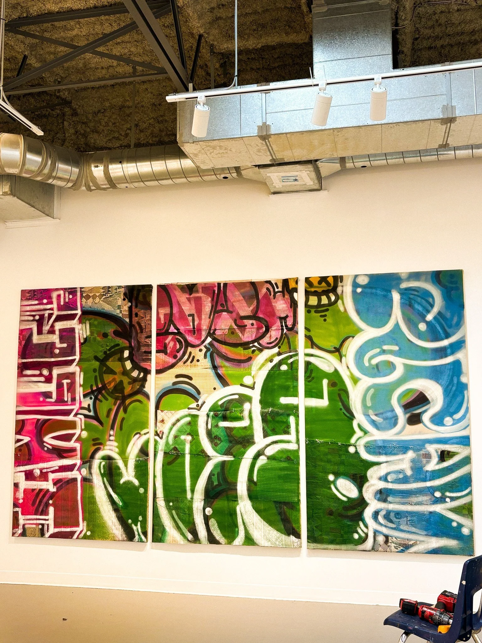 Colorful graffiti art on three panels under an industrial ceiling with exposed ducts and track lighting; a chair with items underneath.