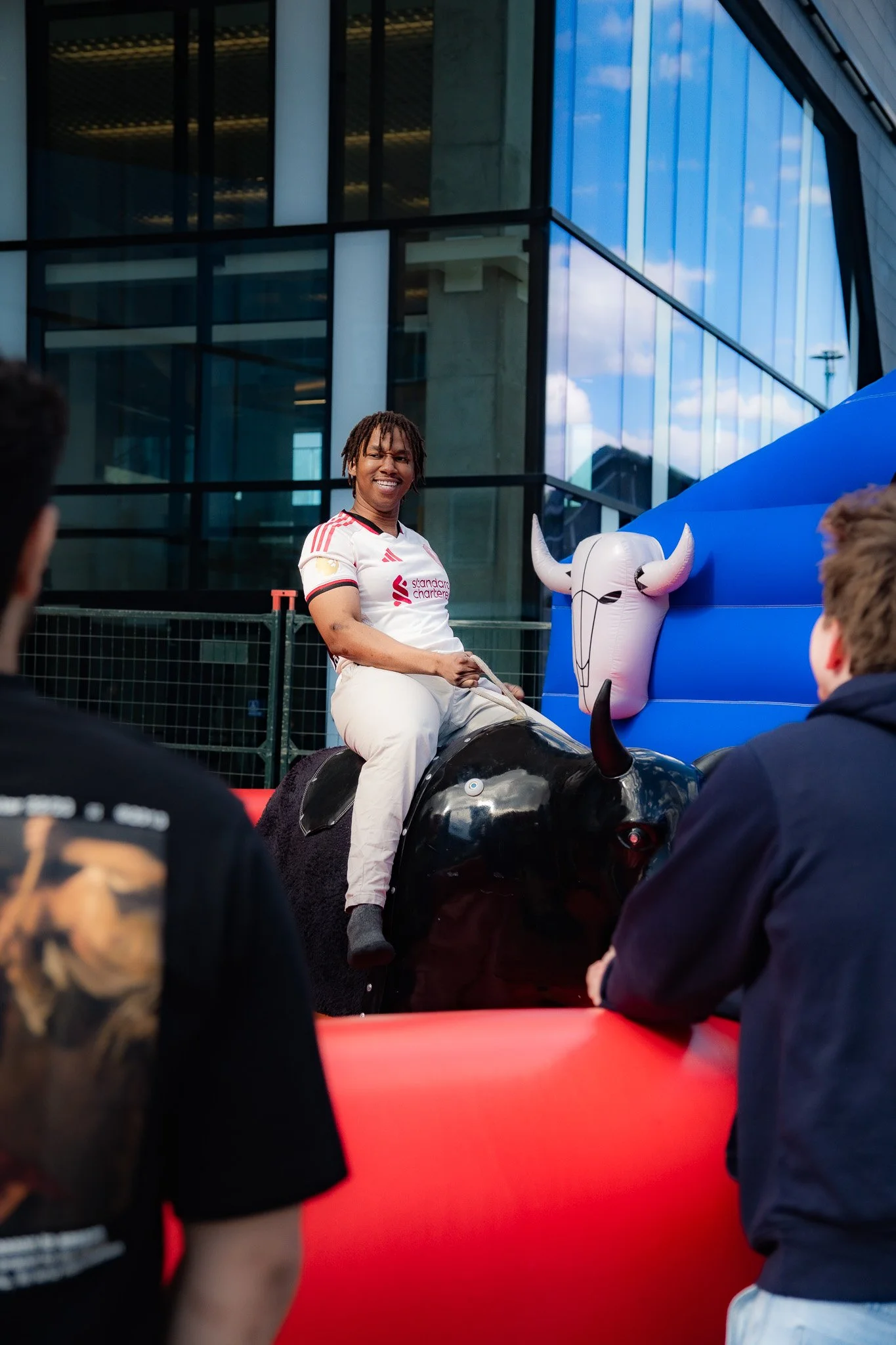 A person wearing a white sports jersey with red stripes and text sitting on a mechanical bull at an outdoor event, with other people around and modern buildings with glass windows in the background.