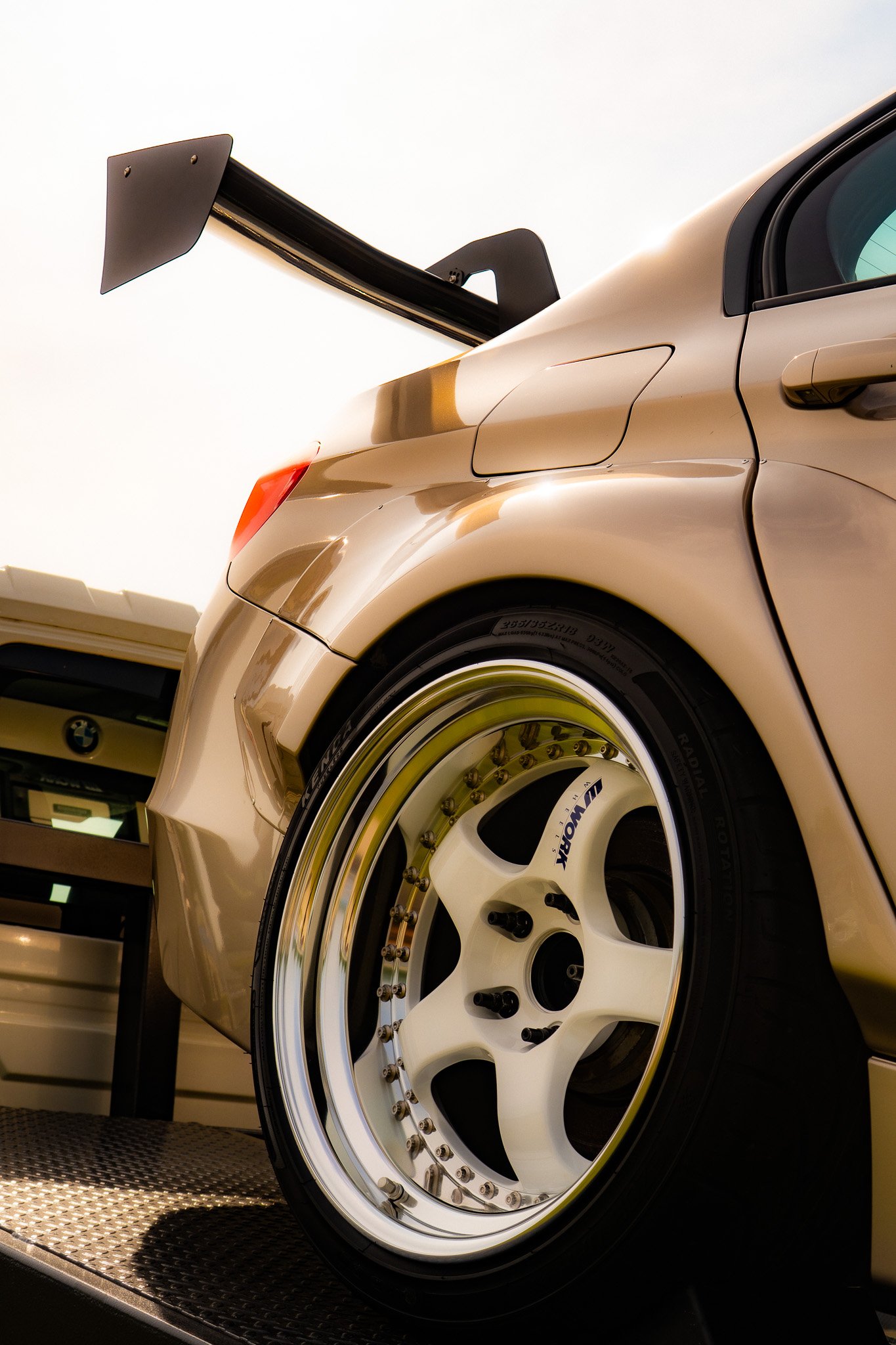 Close-up of a race car's rear wheel and wing, gold-colored body with a white rim and BMW logo on the wheel, parked on a textured surface.