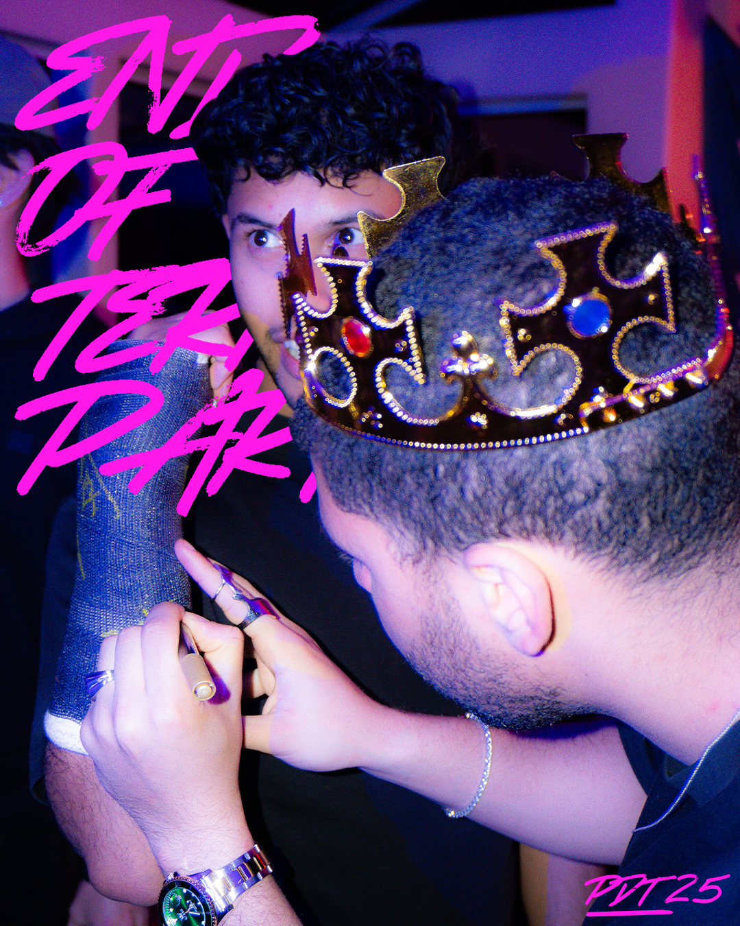 Two young men wearing crowns, indoors in a party setting. One man has curly hair and is looking at the other, who is signing his hand. The photo is overlaid with vibrant pink text that says