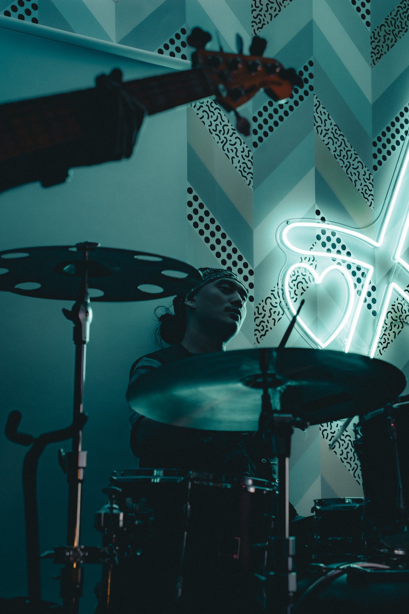 A person playing a drum set in a room with a geometric patterned wall and neon sign in the shape of a heart.