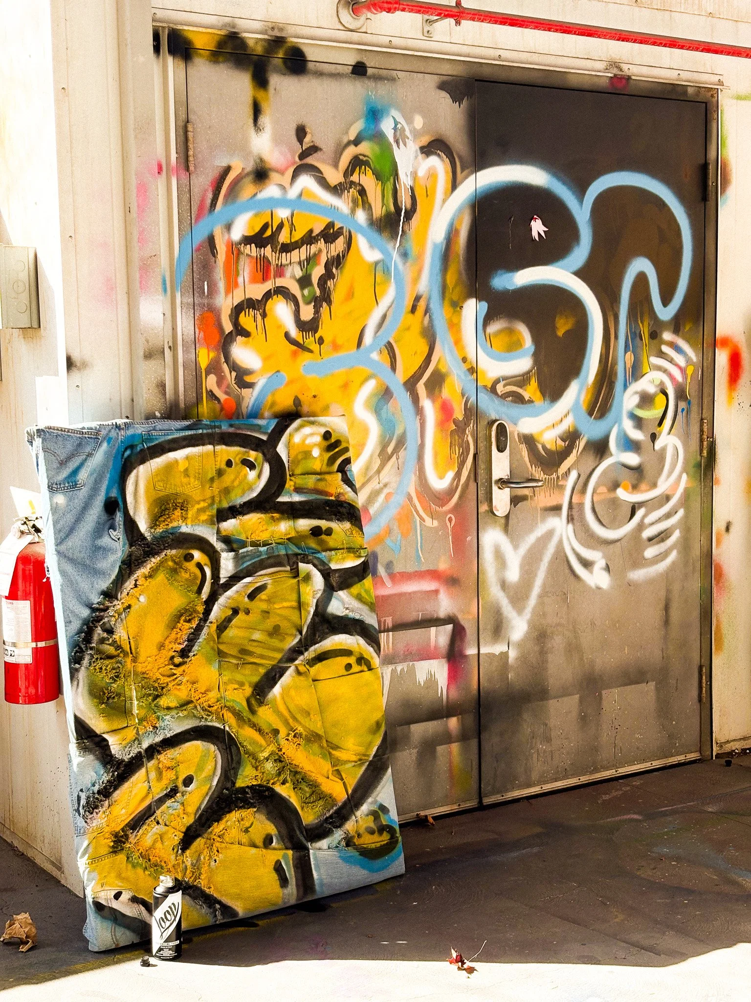 Graffiti art on a metal door and surrounding wall, featuring colorful abstract designs with dominant yellow and blue shapes. A painted canvas with similar artistic elements leans against the wall next to a fire extinguisher and a spray paint can on the ground.