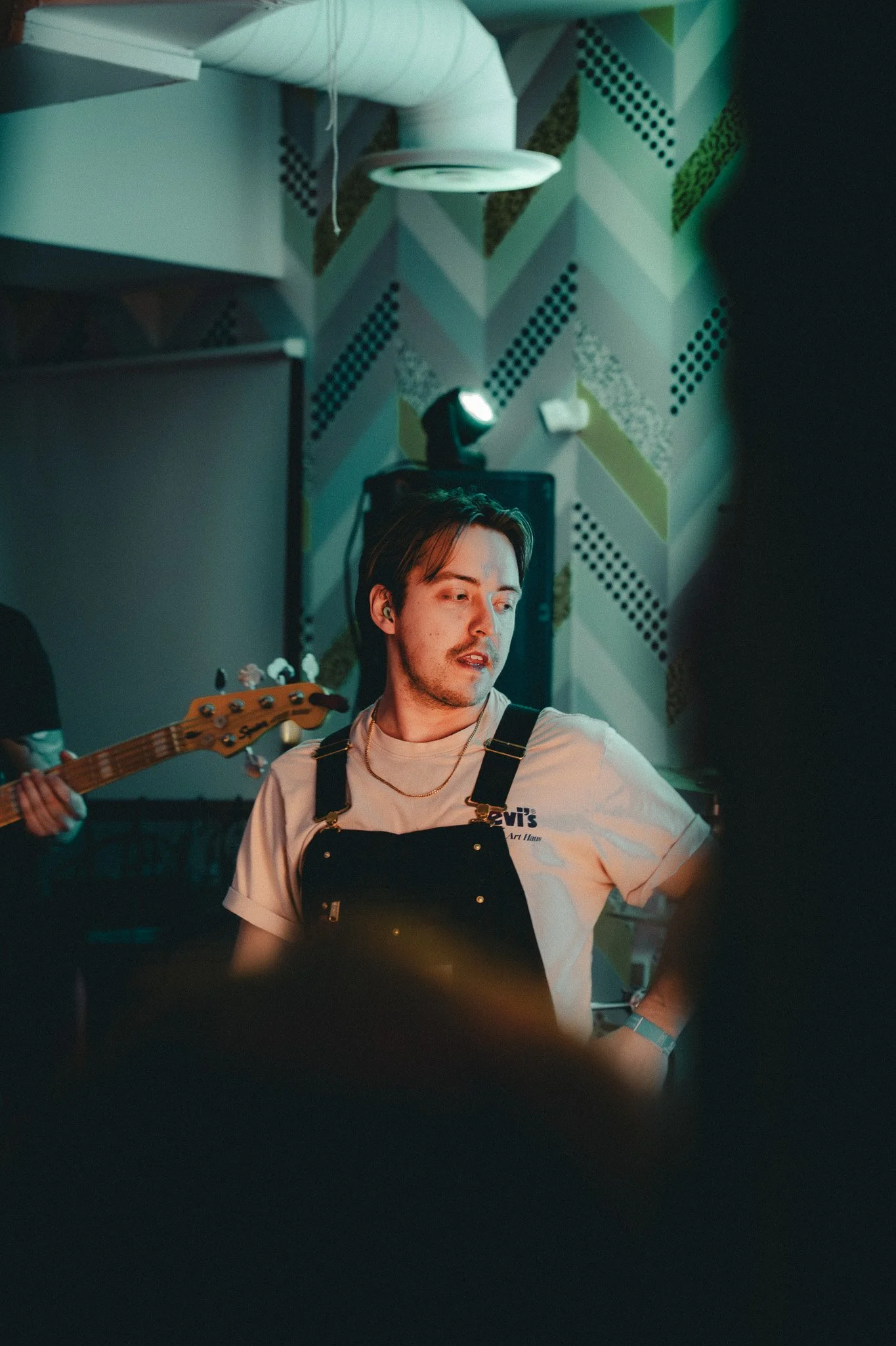A young man with dark hair and facial hair, wearing a beige T-shirt with a Levi's logo, a black apron, and a silver chain necklace, is performing or rehearsing on a stage with colorful, patterned geometric wallpaper in the background. Stage equipment including a spotlight and a guitar are also visible.