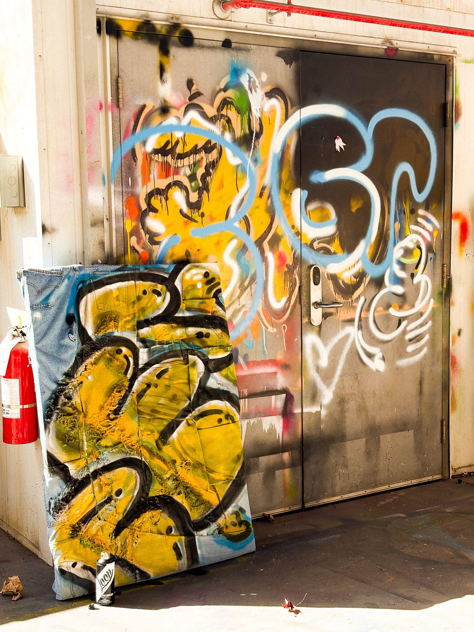 Graffiti on metal door and canvas, featuring colorful abstract designs with prominent yellow and blue elements; a fire extinguisher and spray paint can are visible nearby.