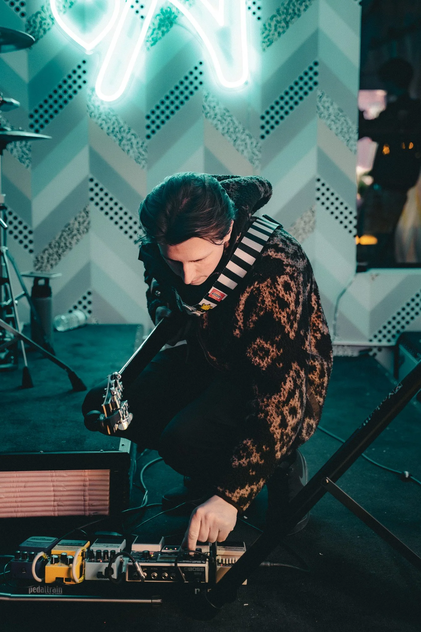 Person with dark hair wearing a patterned jacket, sitting and adjusting a musical instrument connected to various effects pedals on the floor, with a geometric patterned wall and neon sign in the background.