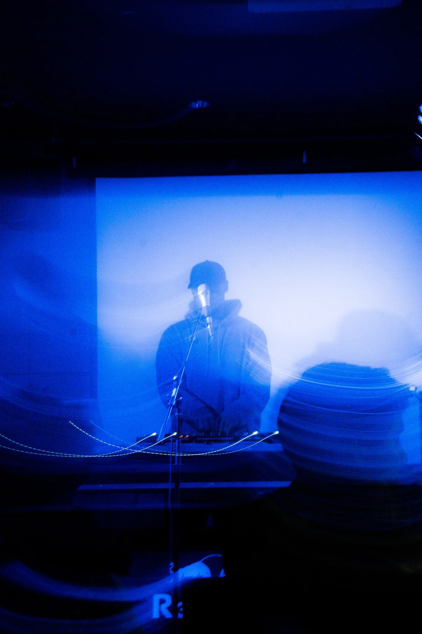 A person speaking into a microphone on a stage with a large screen behind them, illuminated with blue lighting.