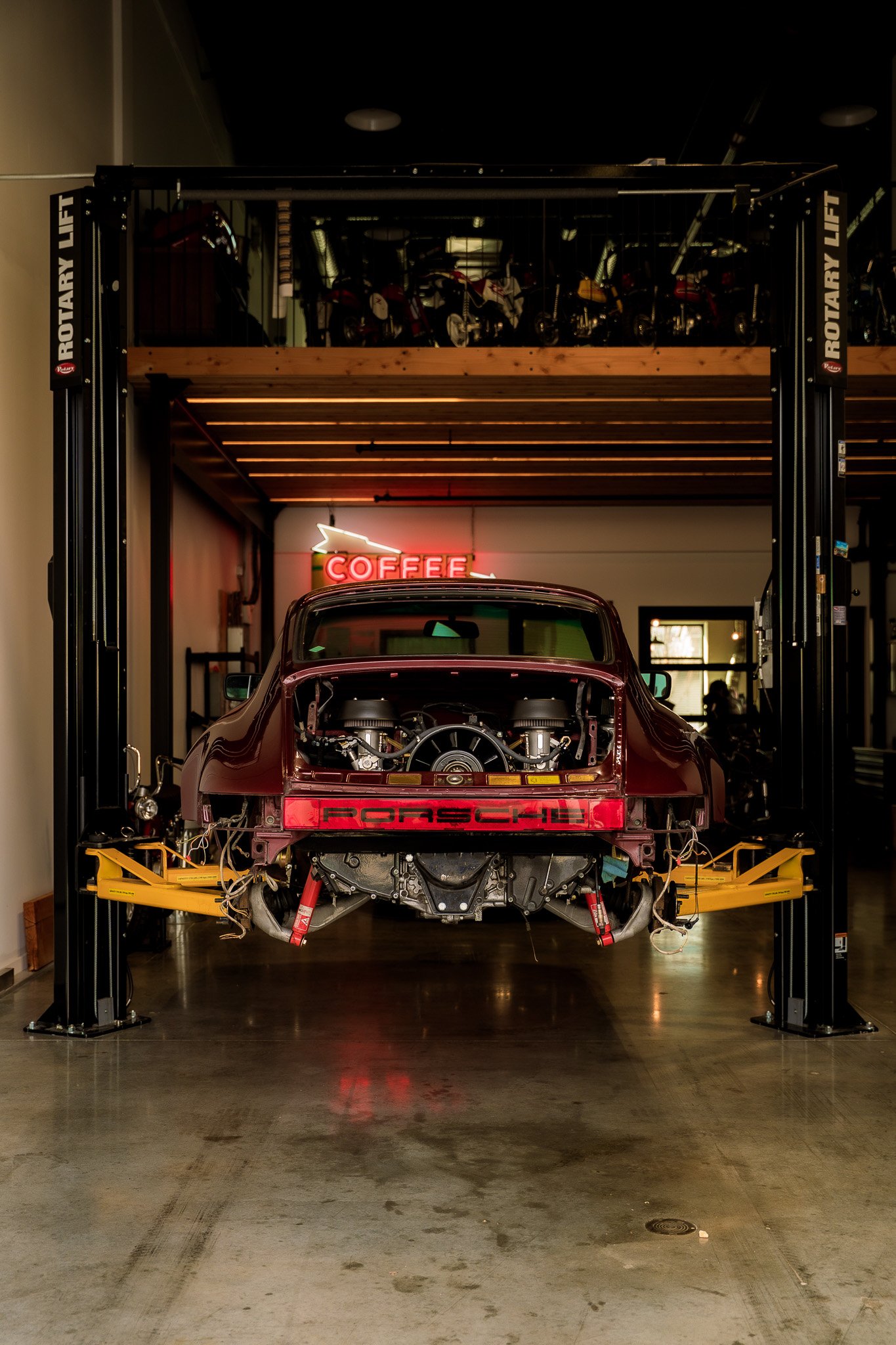 A vintage Porsche car with the engine exposed is on a hydraulic lift inside a garage or workshop, with a neon 'COFFEE' sign on the wall and motorcycles on a mezzanine above.