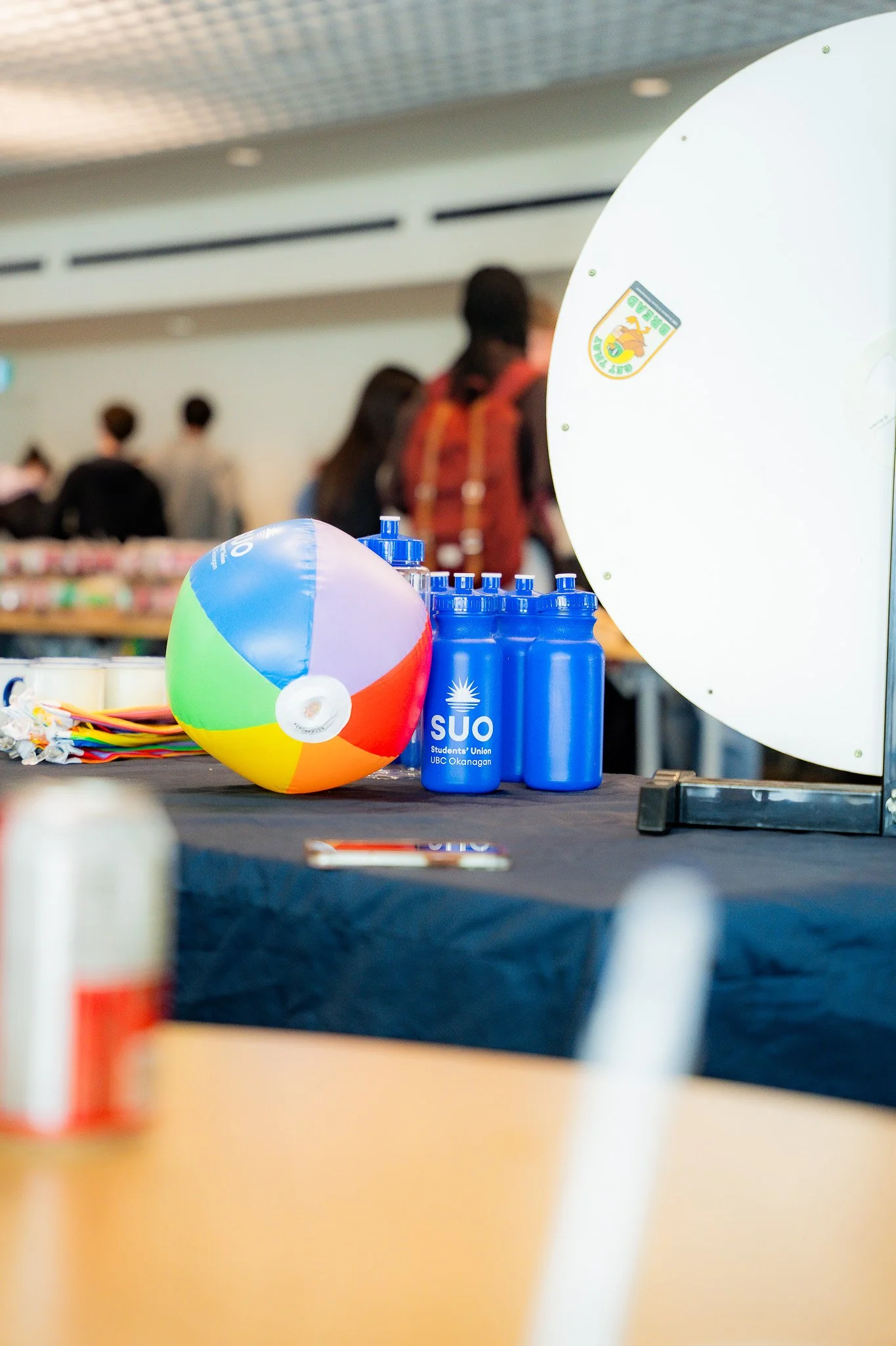 A colorful beach ball, three blue water bottles, and a small orange container are on a table at an indoor event, with blurred people and background in the distance.