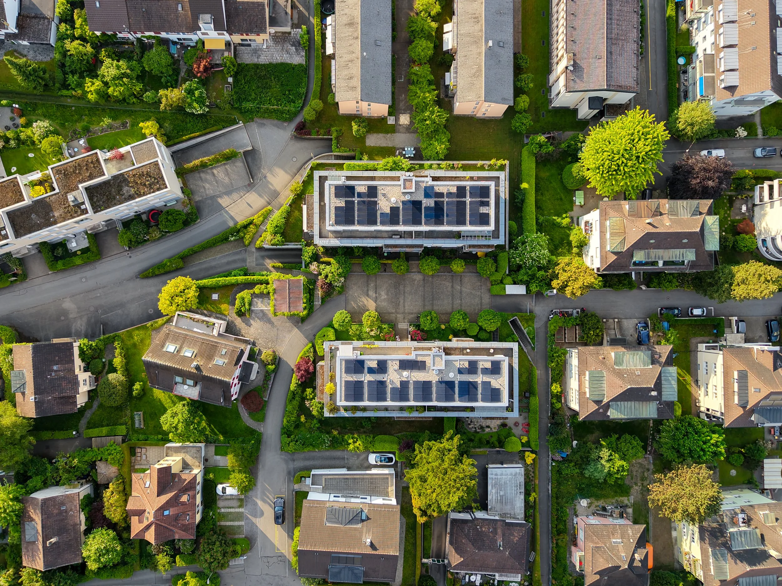 Vogelperspektive eines Wohnquartiers mit mehreren Mehrfamilienhäusern, Gärten, Straßen und parkenden Autos, umgeben von Bäumen und Grünflächen.