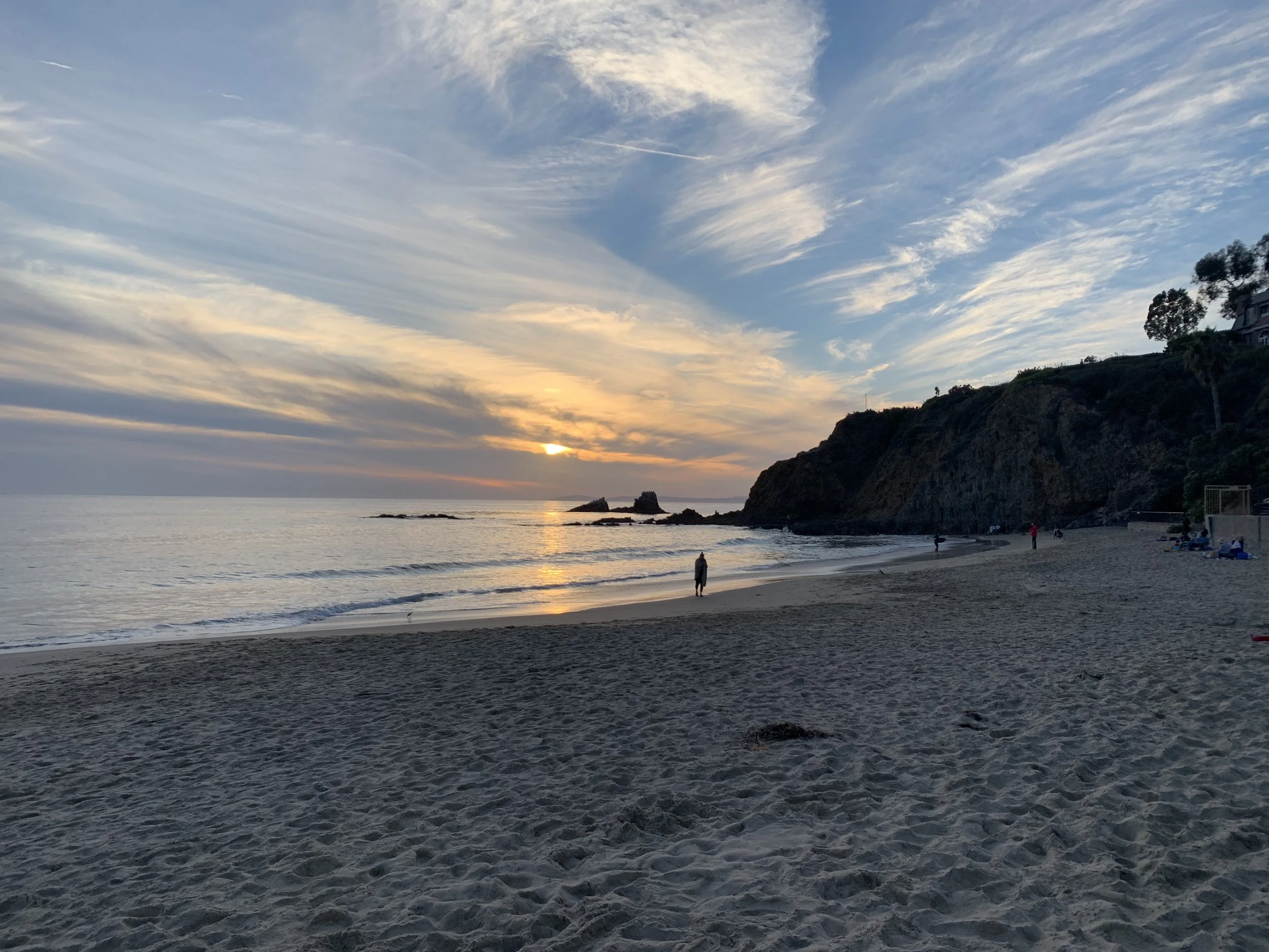 Beach at sunset with cliffs, ocean waves, and a person walking along the shore. Sky filled with clouds and setting sun.