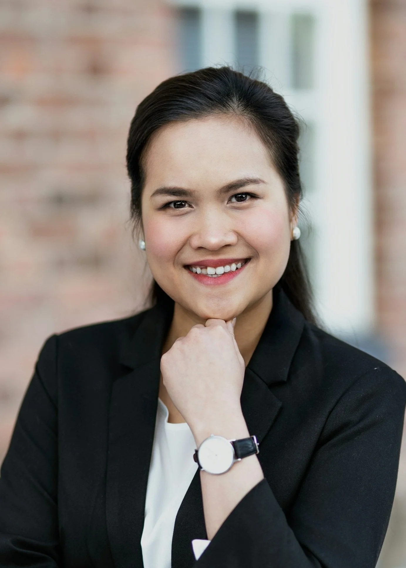 A woman with dark hair in a ponytail, wearing a black blazer, white top, pearl earrings, and a wristwatch, smiling with her hand resting on her chin, standing outdoors with a blurred brick and window background.