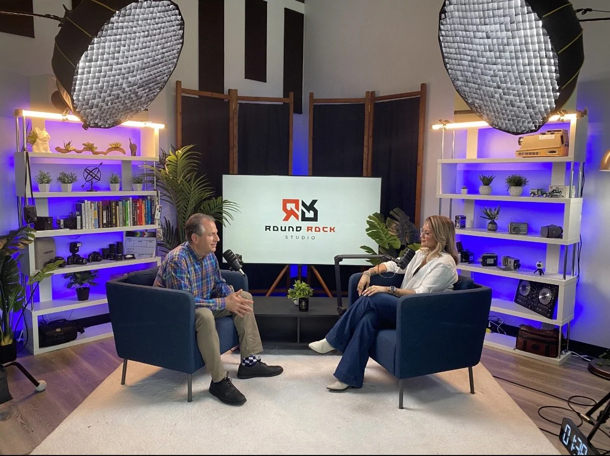 Two people speaking in a recording studio, seated in armchairs, with a large screen behind them displaying the 'Round Rock Studio' logo. The studio has shelves with books, plants, cameras, and audio equipment, illuminated by purple and white lights, and large studio lights overhead.