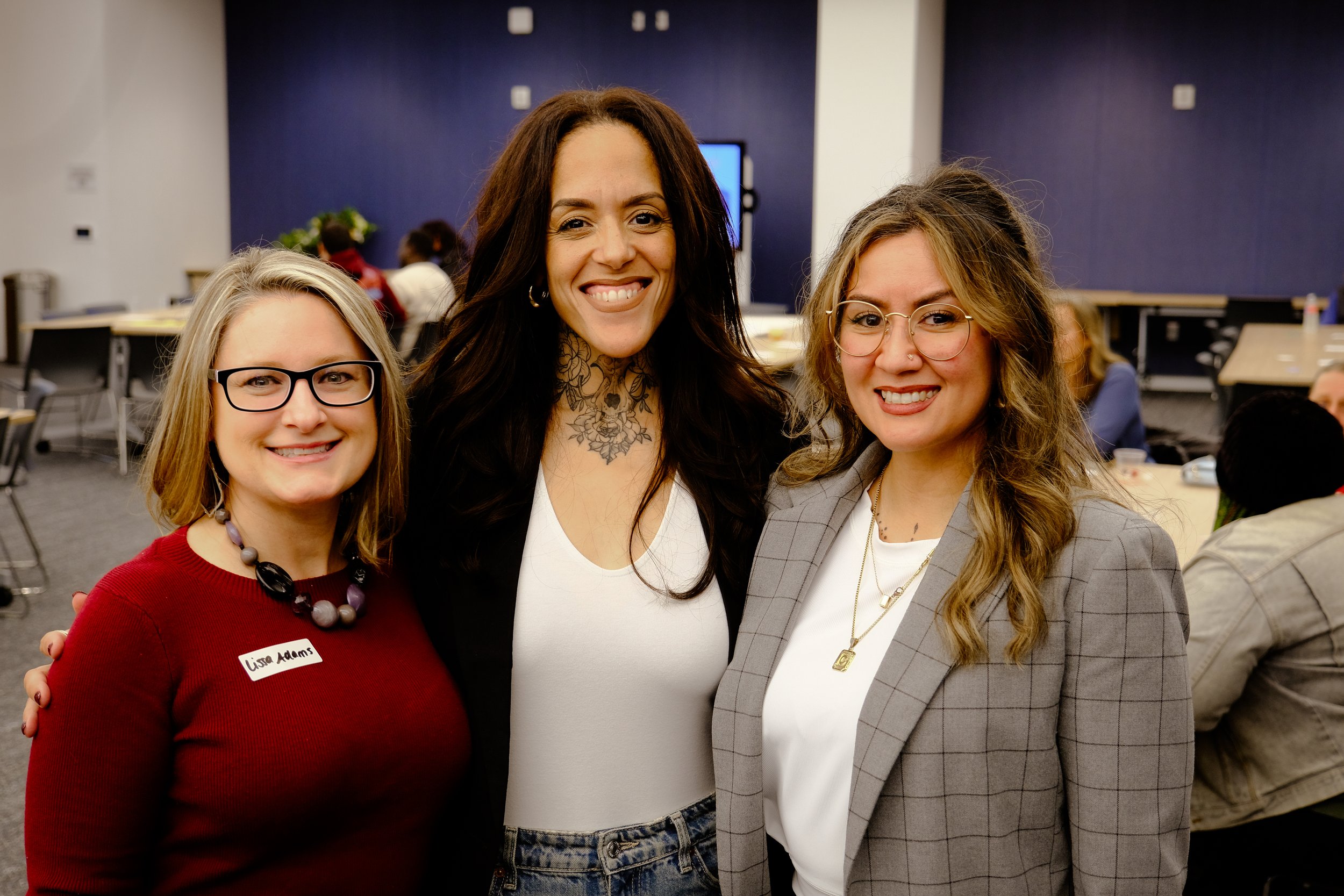 Three women standing together at an indoor event, smiling for the camera, with tables and other people in the background.