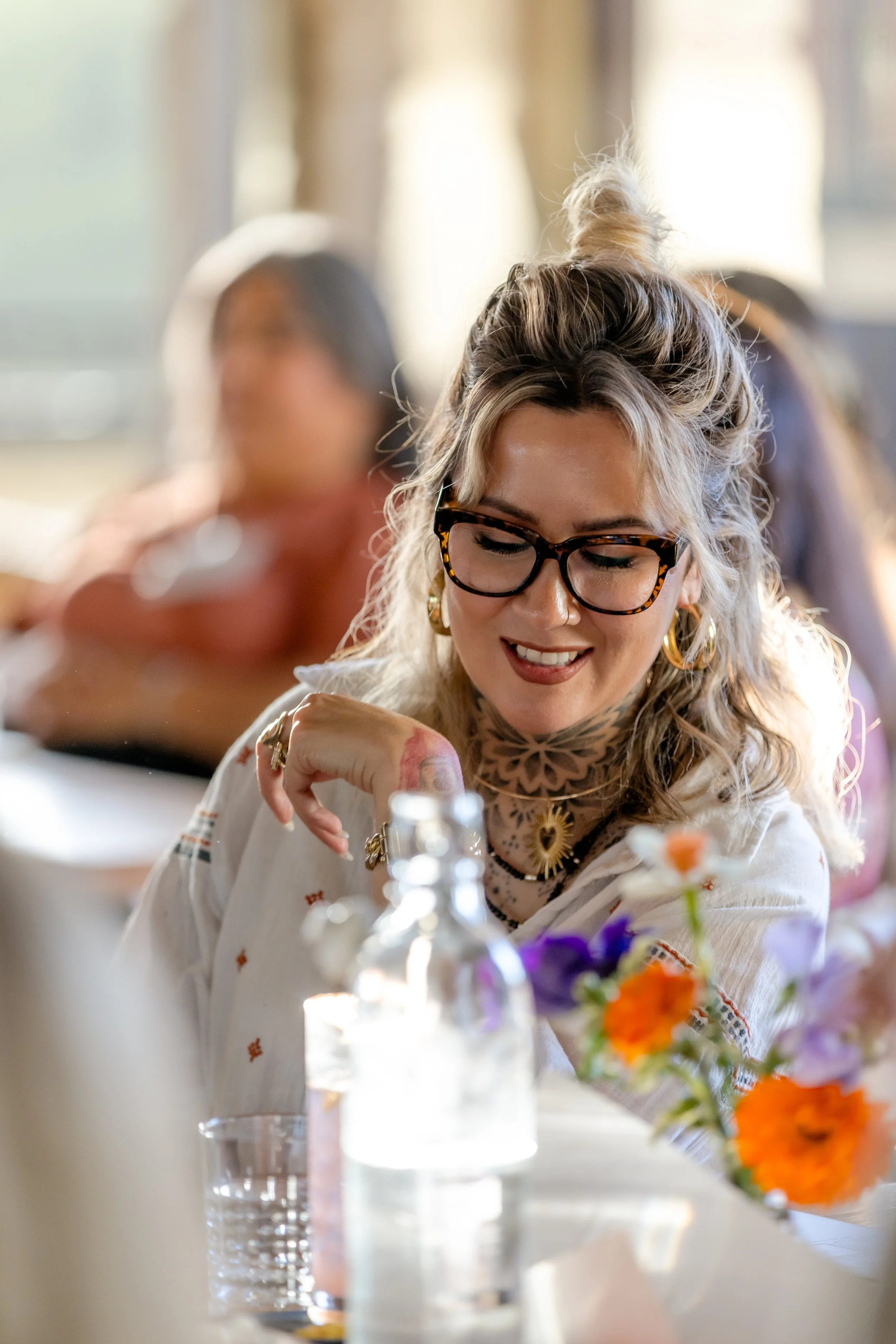 A woman with blonde curly hair, wearing glasses and gold jewelry, smiling at a table with a colorful flower arrangement and bottles, in a well-lit indoor setting.