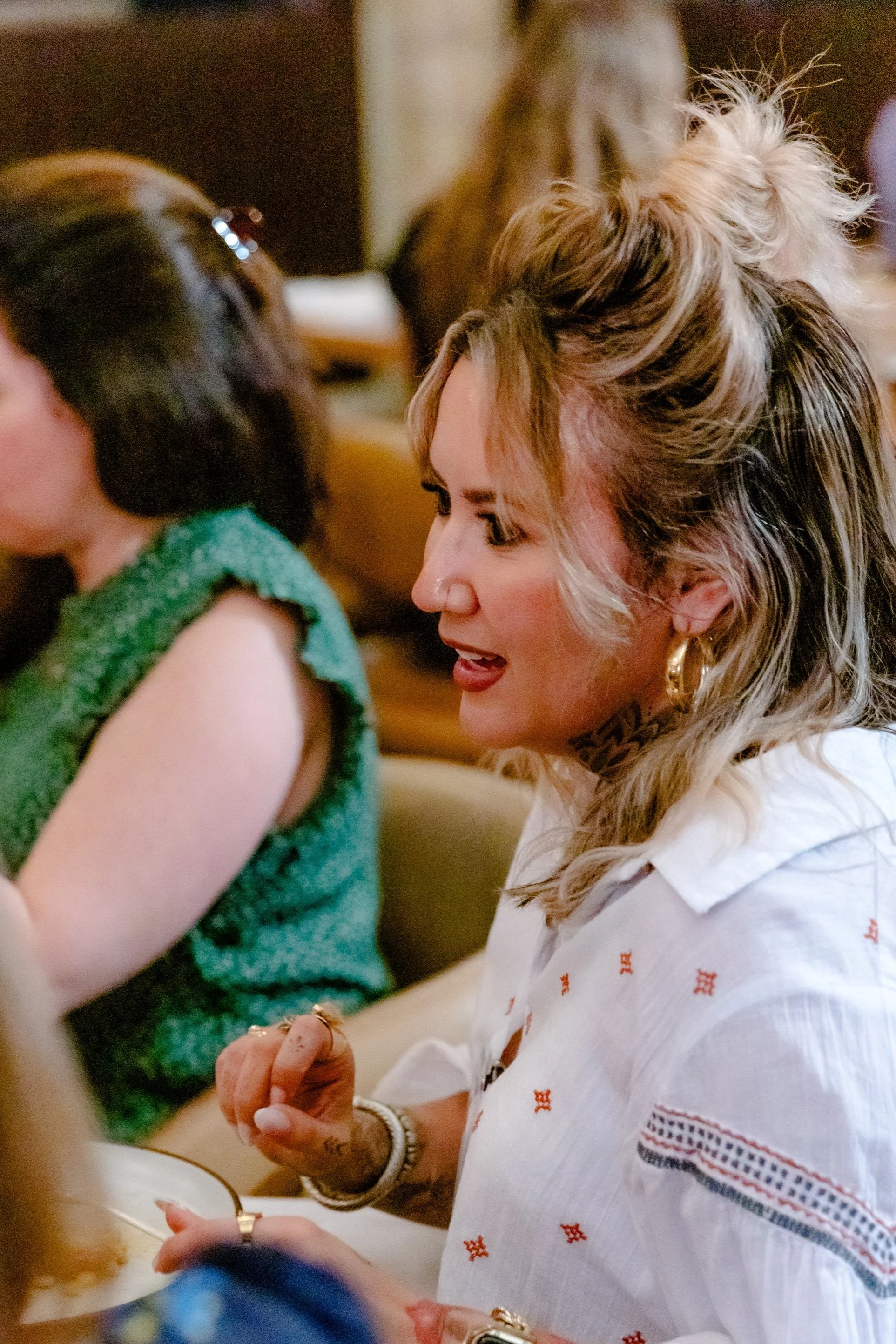 A woman with blonde hair styled in a messy bun, wearing large hoop earrings and a white embroidered shirt, smiling and engaging in conversation at a gathering.