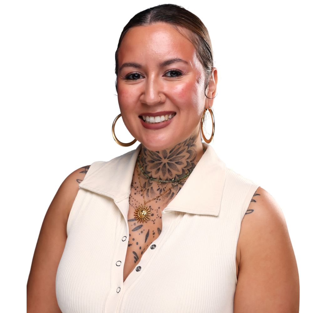 A woman with tattoos on her neck and shoulders, wearing a sleeveless white top, gold hoop earrings, and layered necklaces, smiling at the camera.