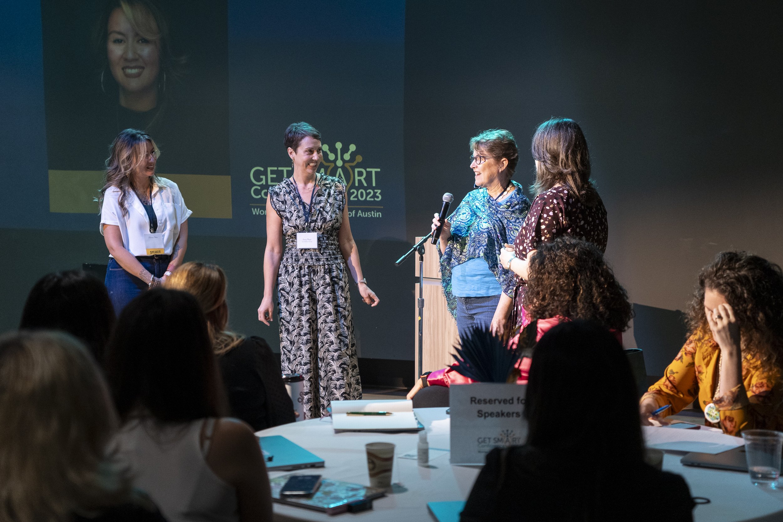 Four women on stage at a conference, one speaking into a microphone, with a large screen displaying a woman’s portrait and the text 'GET SMART Conference 2023' in the background. Audience members are seated at tables.