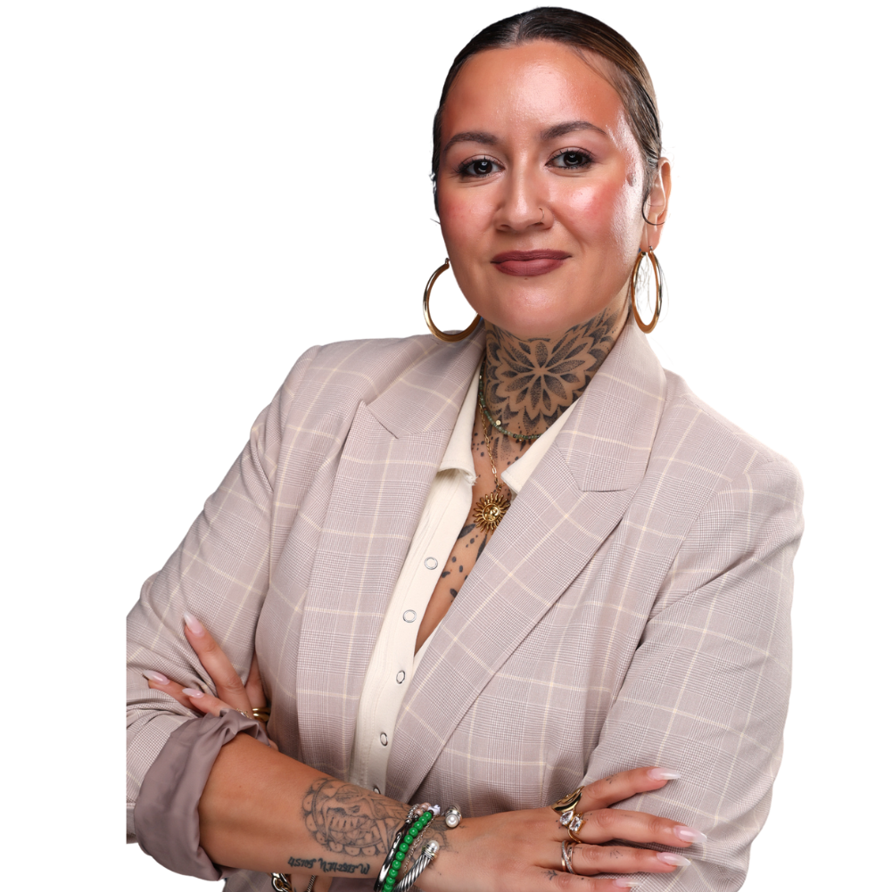 A woman with tattoos on her neck and arms, wearing a light beige checkered blazer over a white shirt, with large hoop earrings, multiple rings, and layered necklaces, posing with her arms crossed.