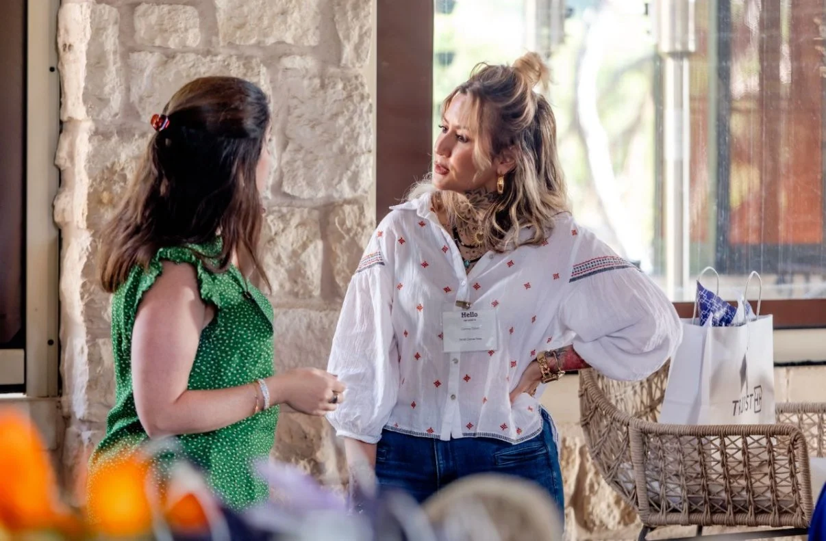 Two women are engaged in a conversation indoors near a stone wall and window, with shopping bags and a wicker chair nearby.