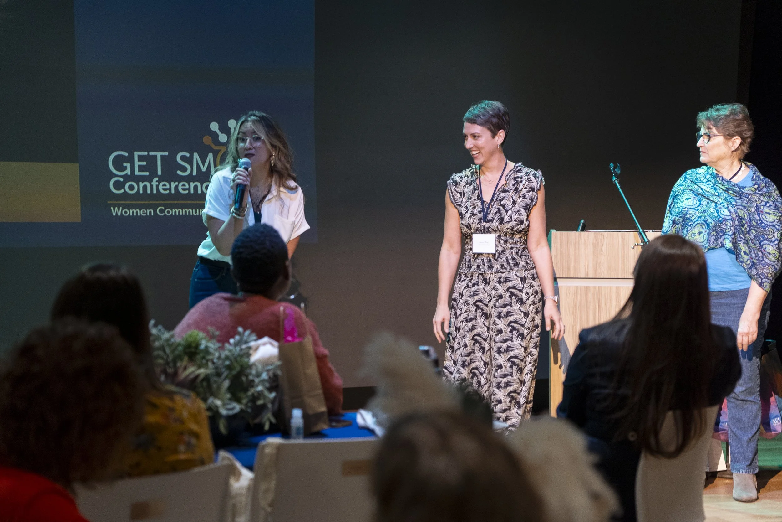 Three women are on a stage at a conference, speaking to an audience. One woman is holding a microphone and speaking, while the other two stand nearby. Audience members are seated at tables facing the stage. There is a large screen behind them displaying the conference logo and title.