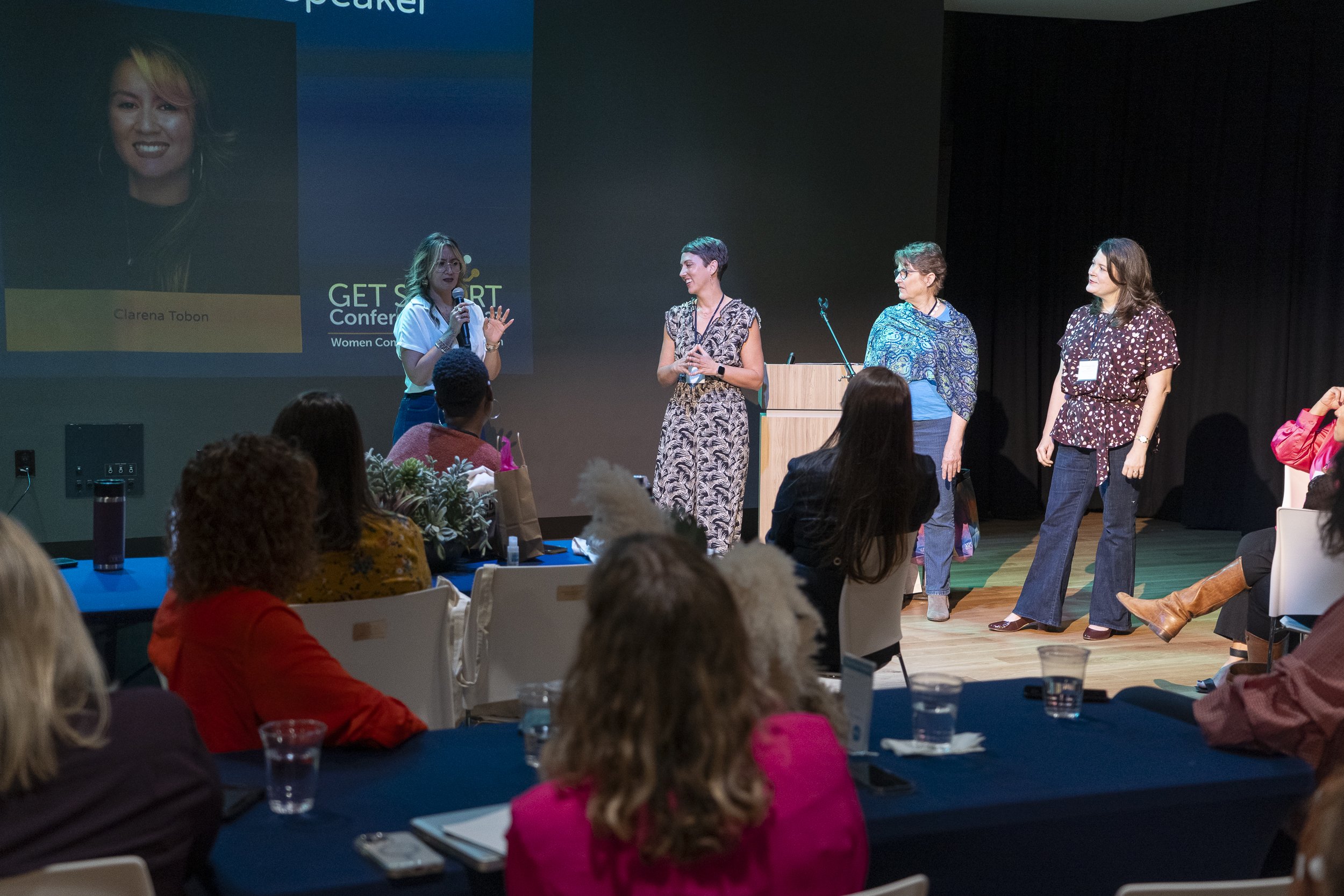 Women speaking on stage at a conference, with an audience seated in front. The backdrop displays the name Clarena Tobon and conference branding.