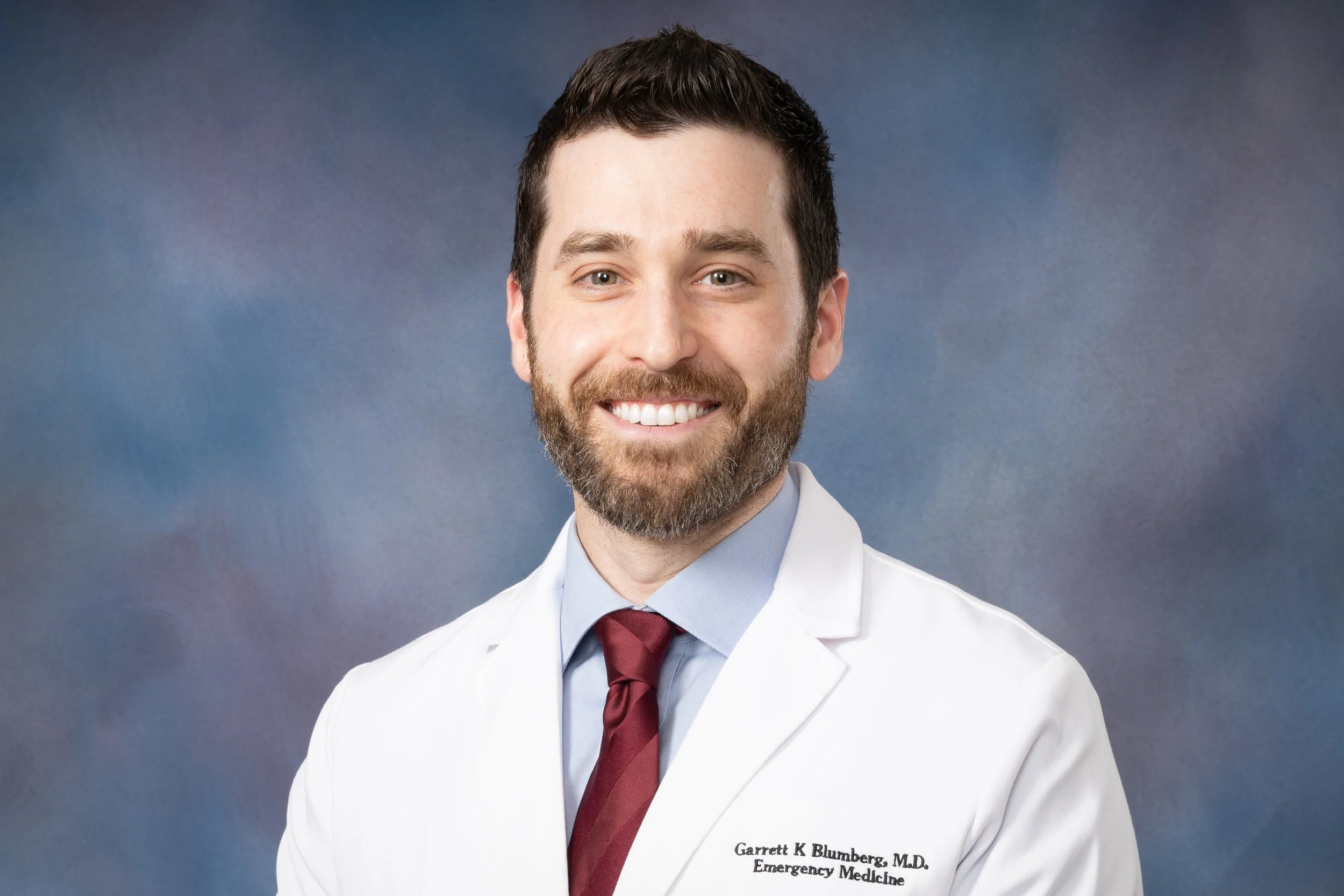 Portrait of a male emergency medicine, emergency room, doctor who provides expert witness services and opinions. He has a beard, looks experienced, is wearing a white coat (lab coat) and red tie, smiling, against a blue gradient background.