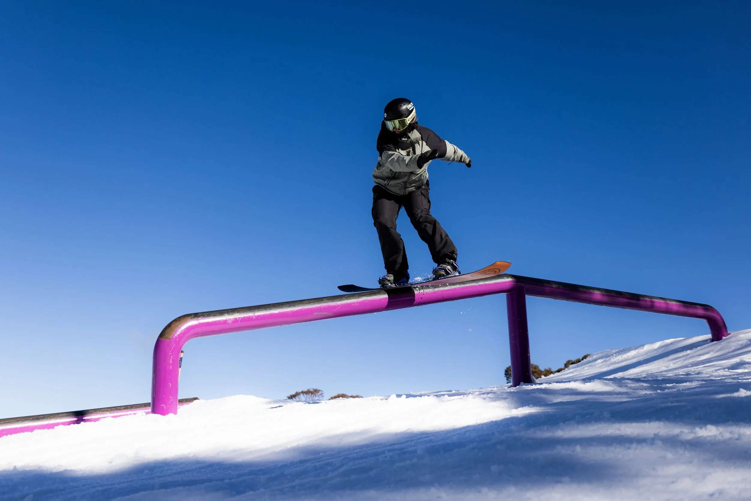 A girl snowboarding on a rail in a snowy landscape under a clear blue sky.