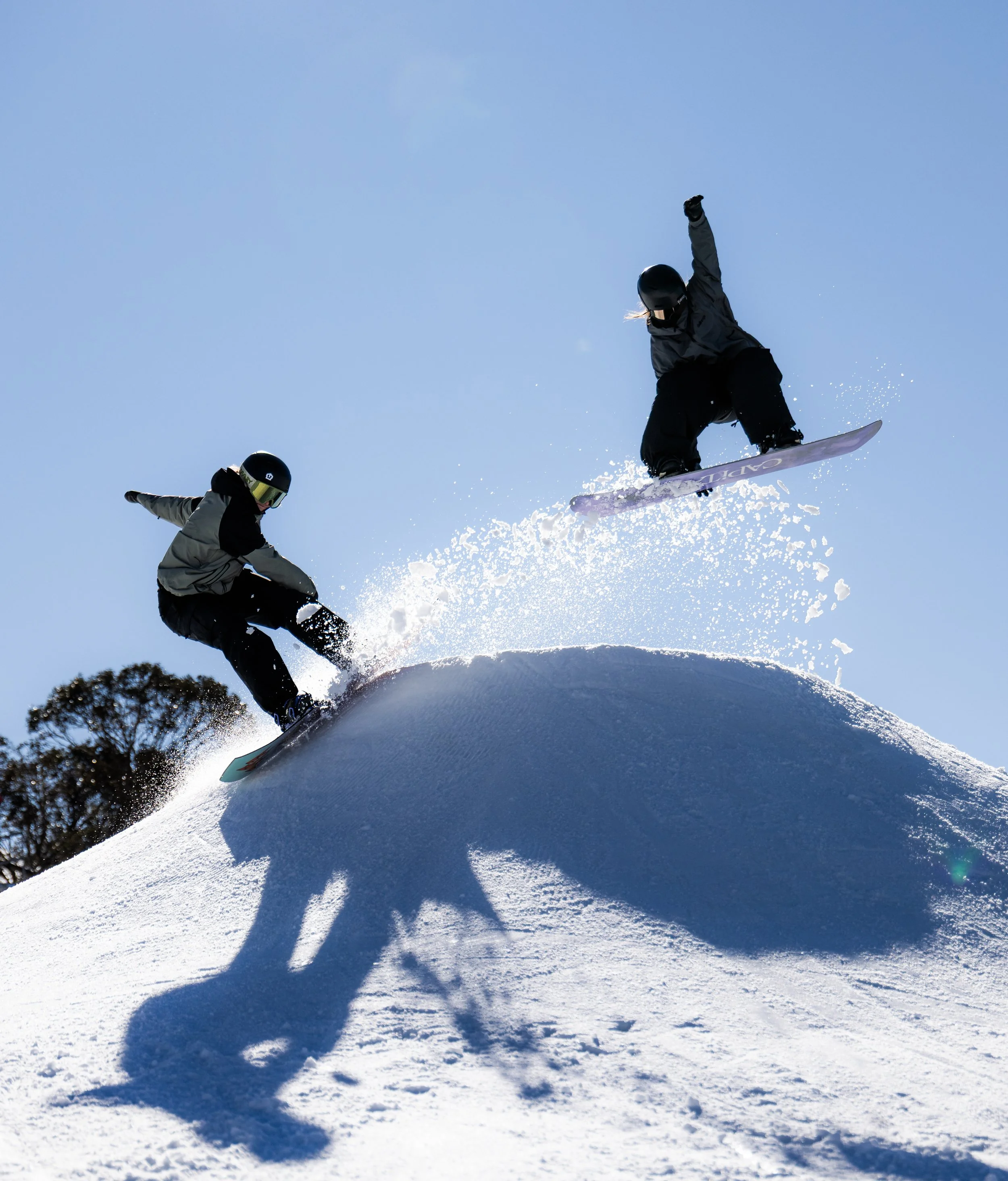 Two girl snowboarders jumping and performing tricks on a snow-covered slope against a clear blue sky.