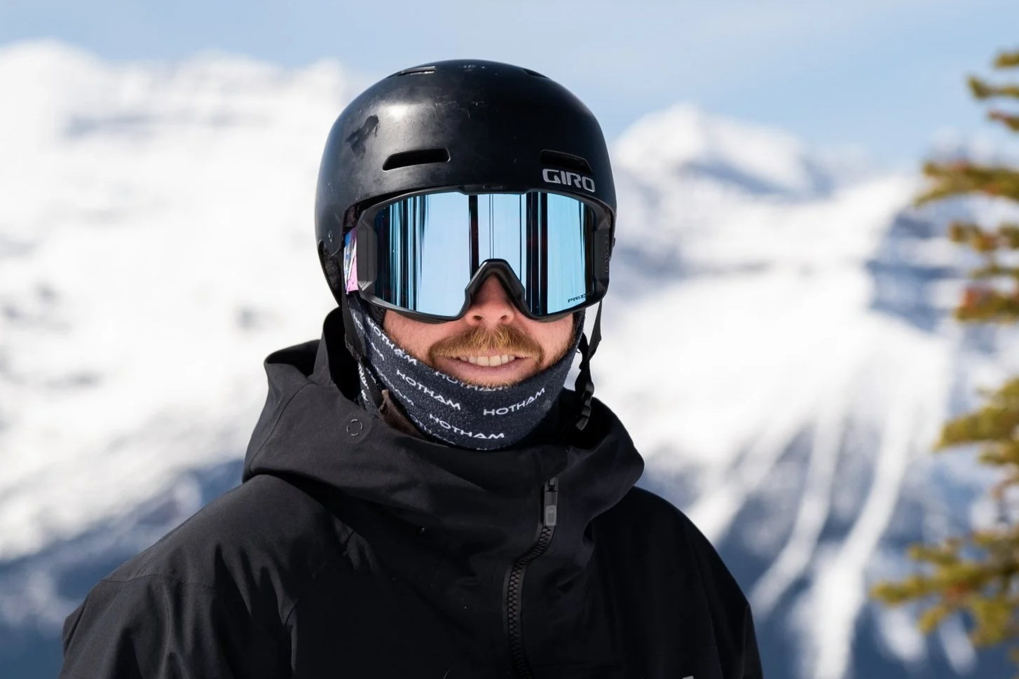A man smiling outdoors with snowy mountains and trees in the background.