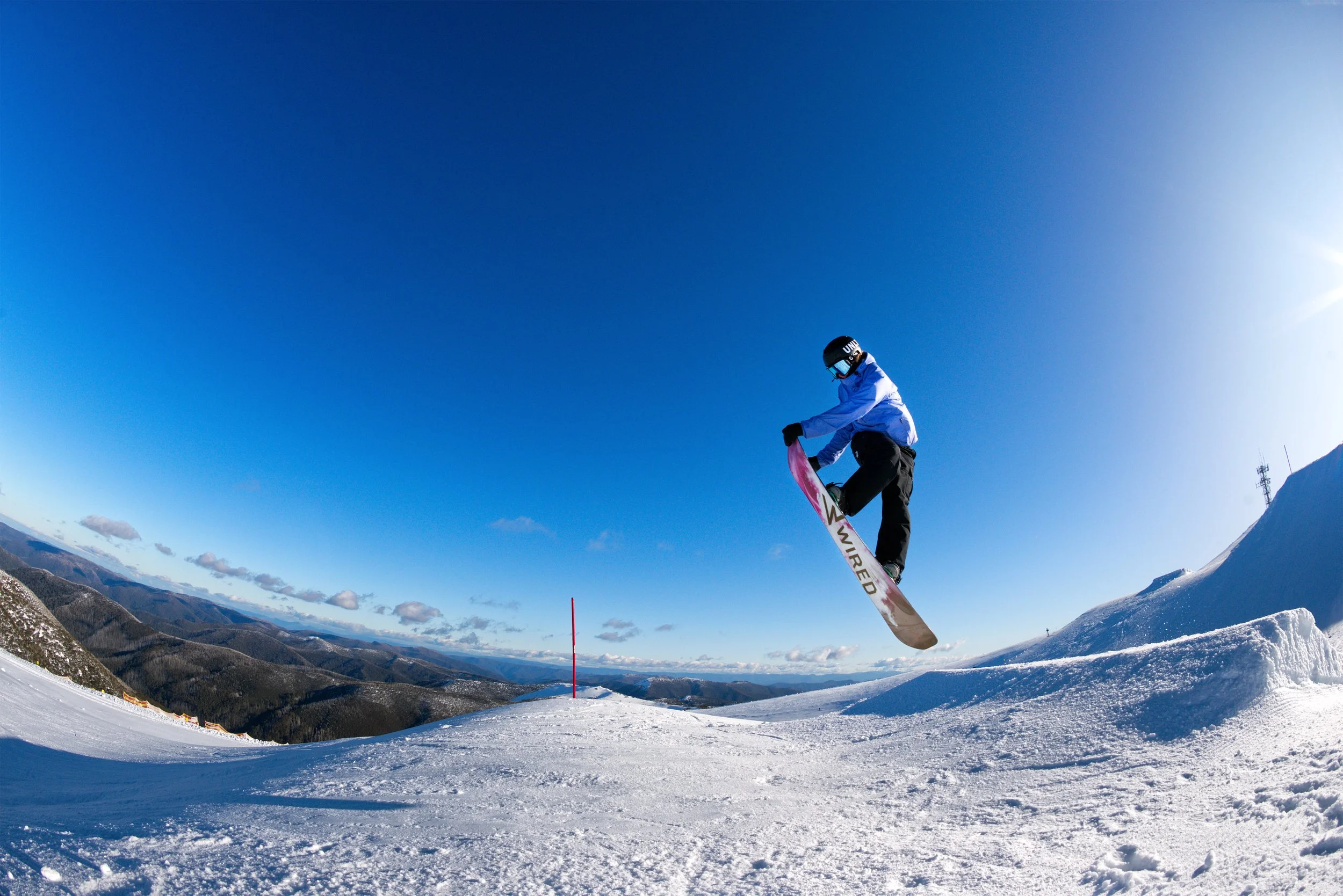 A snowboarder in mid-air performing a jump on a snowy mountain slope during sunny weather, with a clear blue sky and distant mountain ranges in the background.