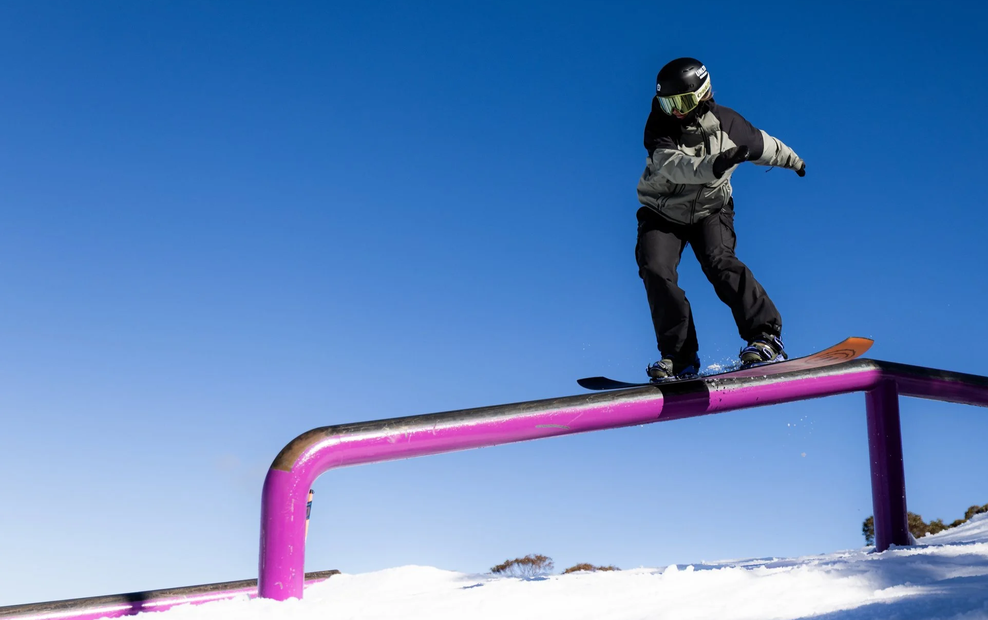 A person snowboarding on a rail in a snow-covered outdoor area under a clear blue sky.