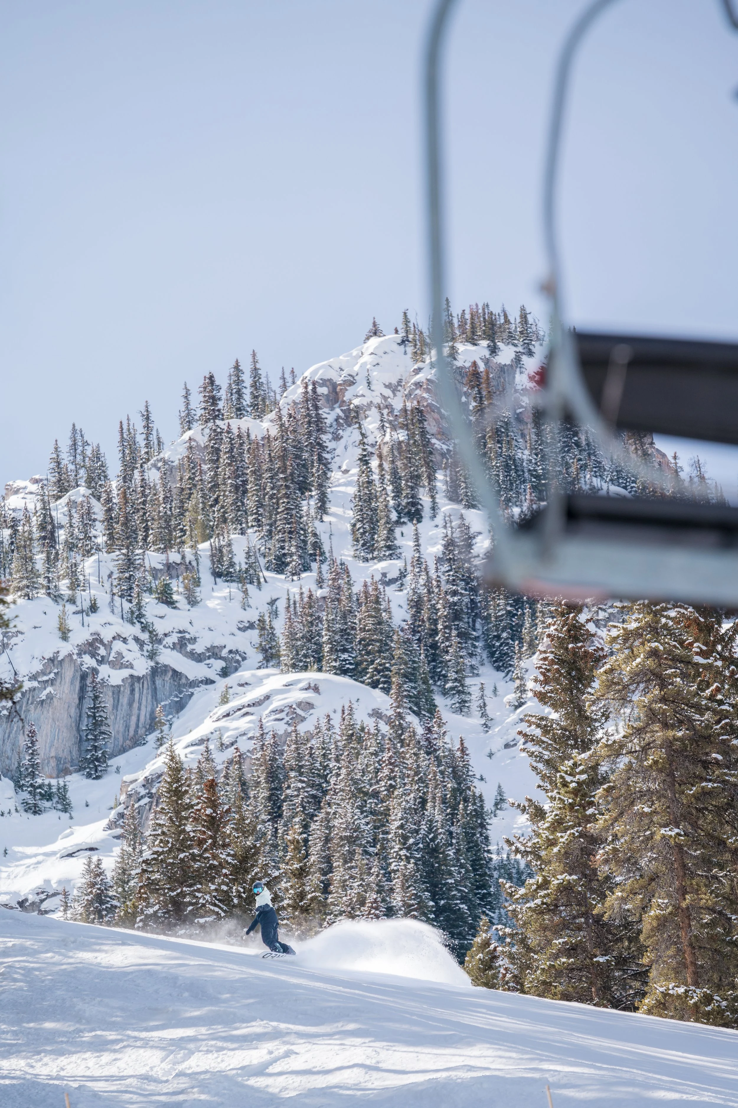 A skier in a snowy mountain landscape with trees and rocky peaks in the background, viewed through a ski lift.