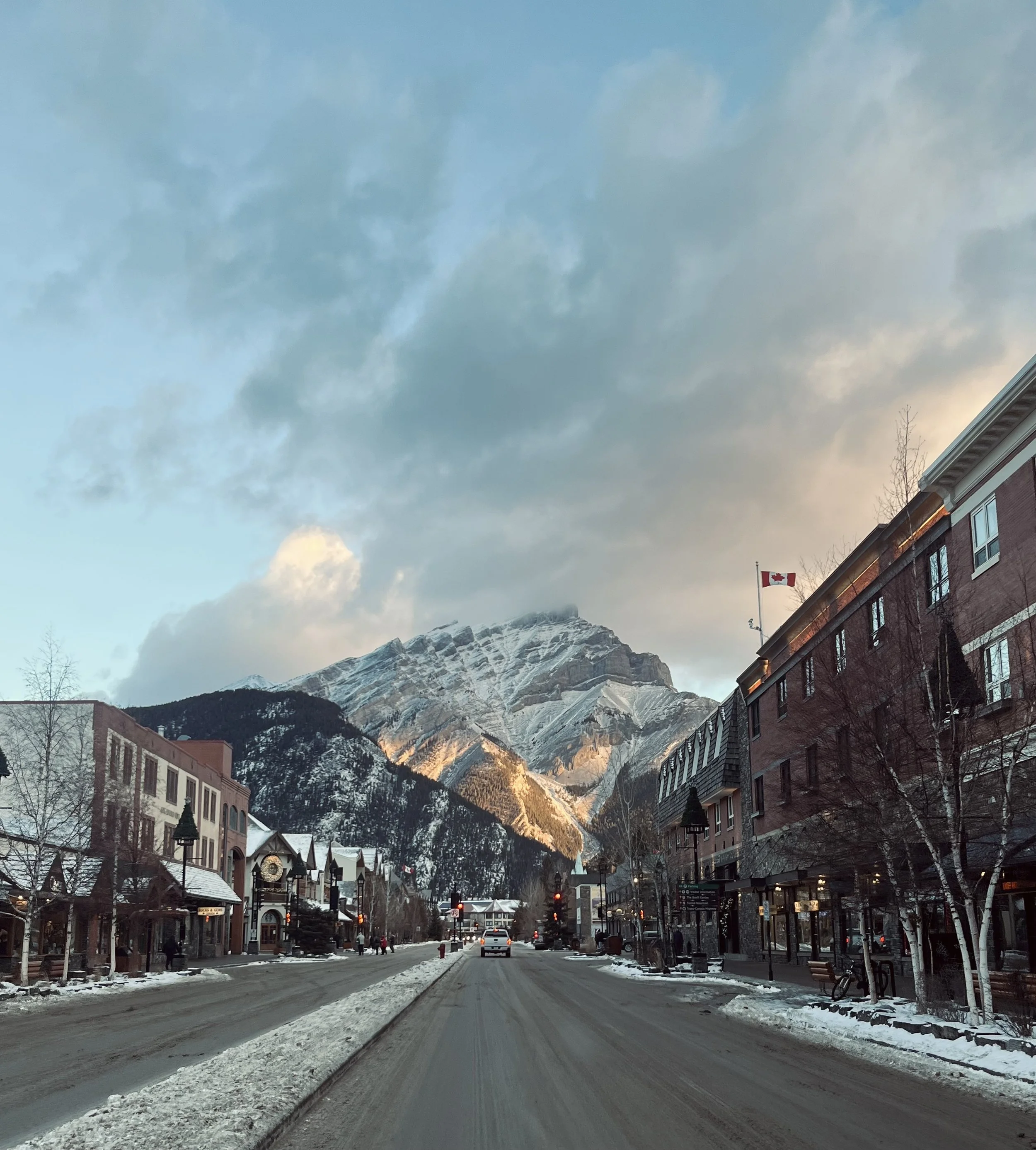 Snow-covered street in a mountain town with a mountain range in the background, storefronts on both sides, and a Canadian flag flying on a building.