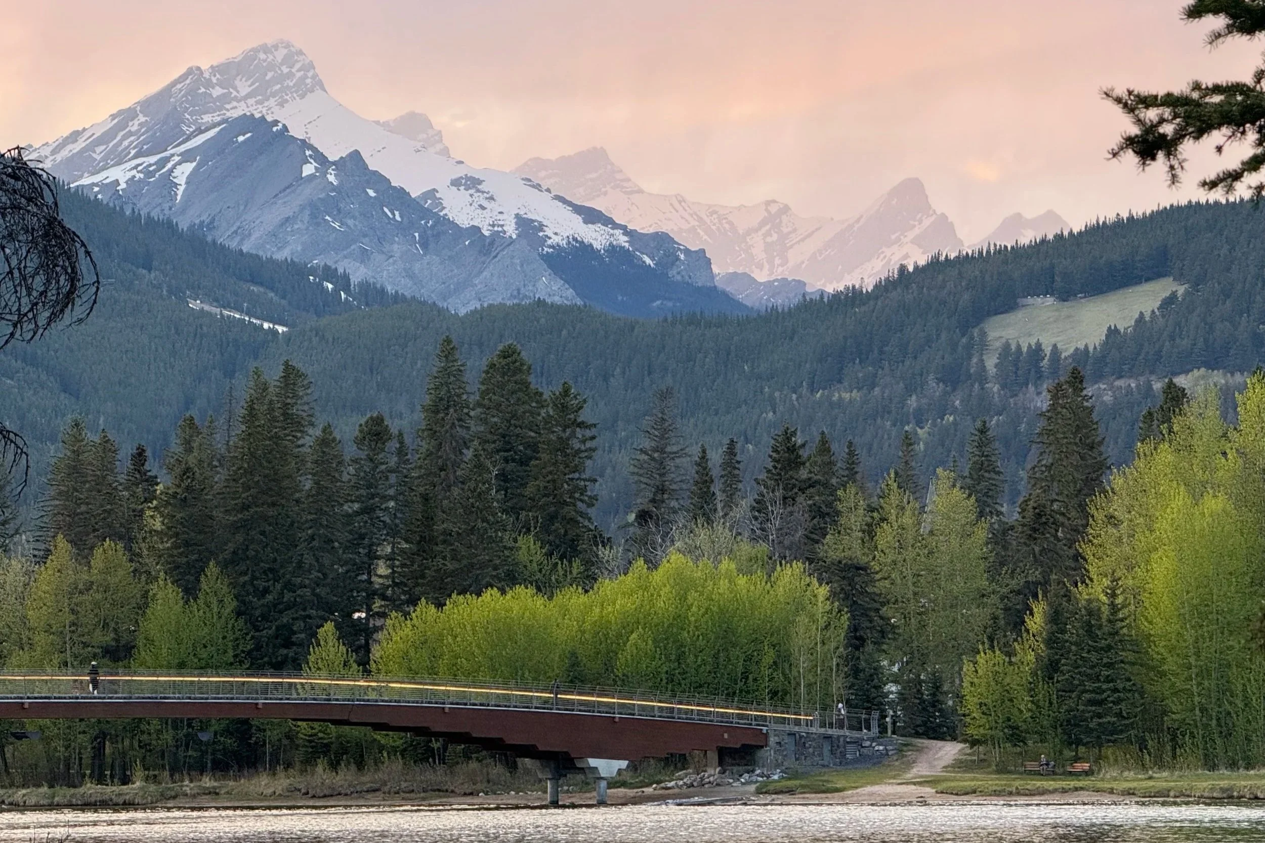 Scenic view of snow-capped mountains, green pine trees, a bridge over a river, and a dirt path in a forested area during daytime.