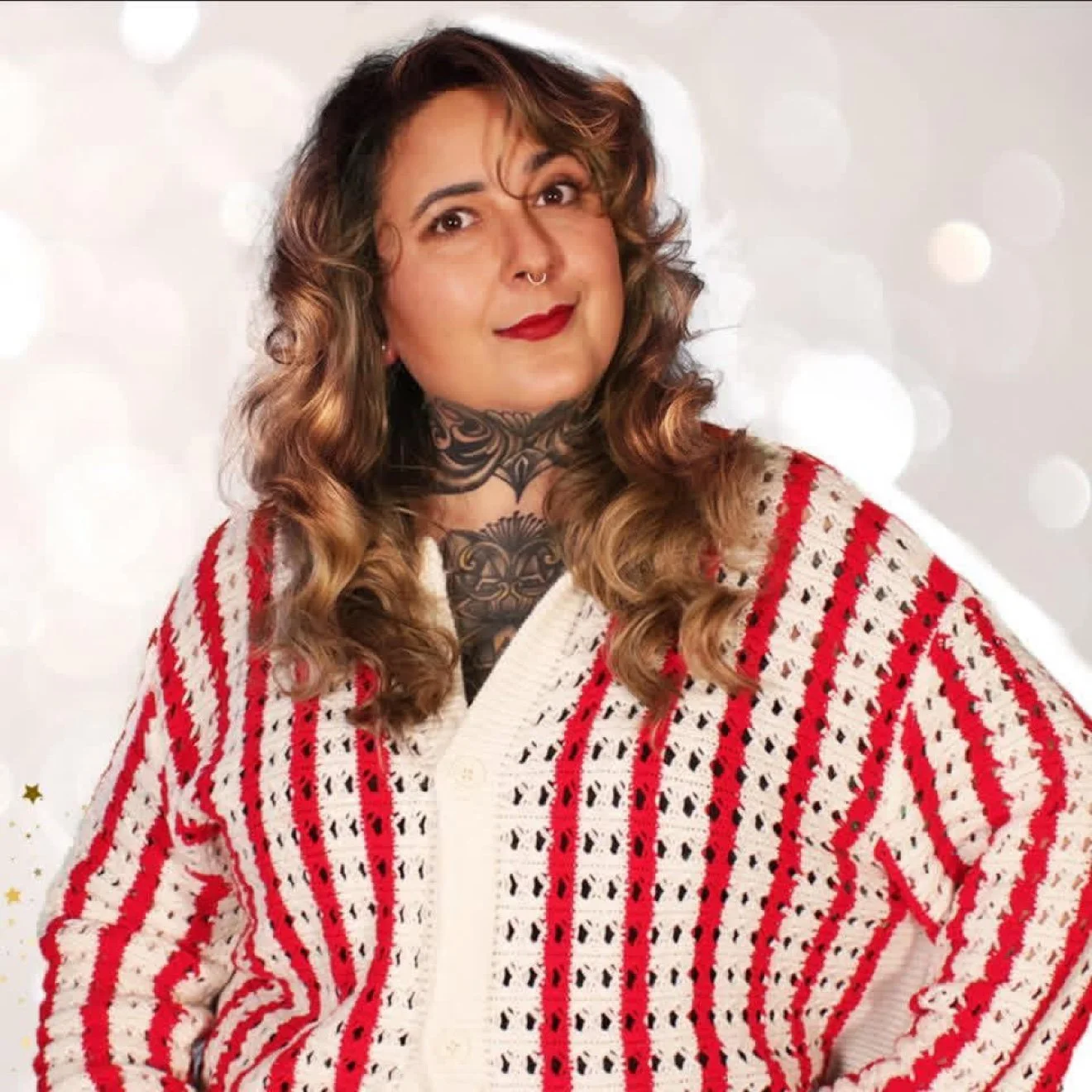 Portrait of a woman with wavy brown hair, tattooed neck, and wearing a red and white patterned cardigan.