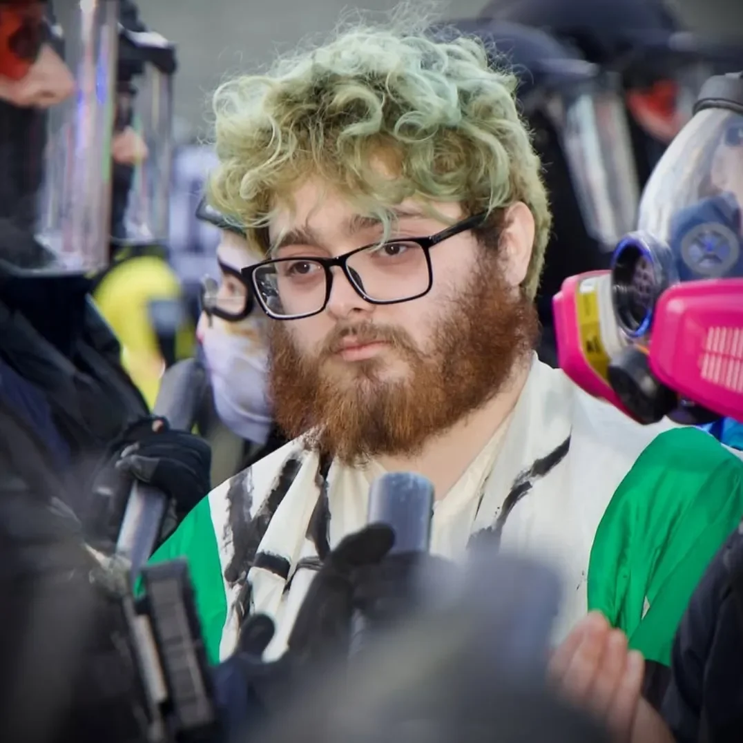 A man with curly blonde hair, glasses, and a beard being interviewed or questioned by multiple people, with police officers and riot gear visible in the background.