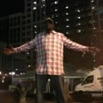 Person standing on a city street at night with arms outstretched, tall buildings illuminated in the background.