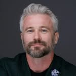 Portrait of a middle-aged man with gray hair and beard, wearing a dark shirt, against a dark background.