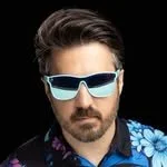 A man with short brown hair and facial hair wearing reflective blue sunglasses and a black shirt with a blue floral pattern.