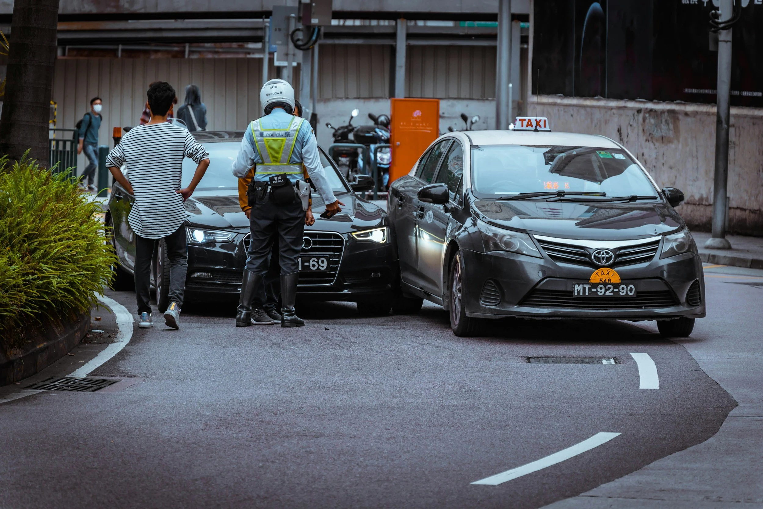A traffic incident involving two cars, a black Audi and a gray Toyota taxi, with a traffic officer assisting in resolving the situation. Two people are standing nearby observing.