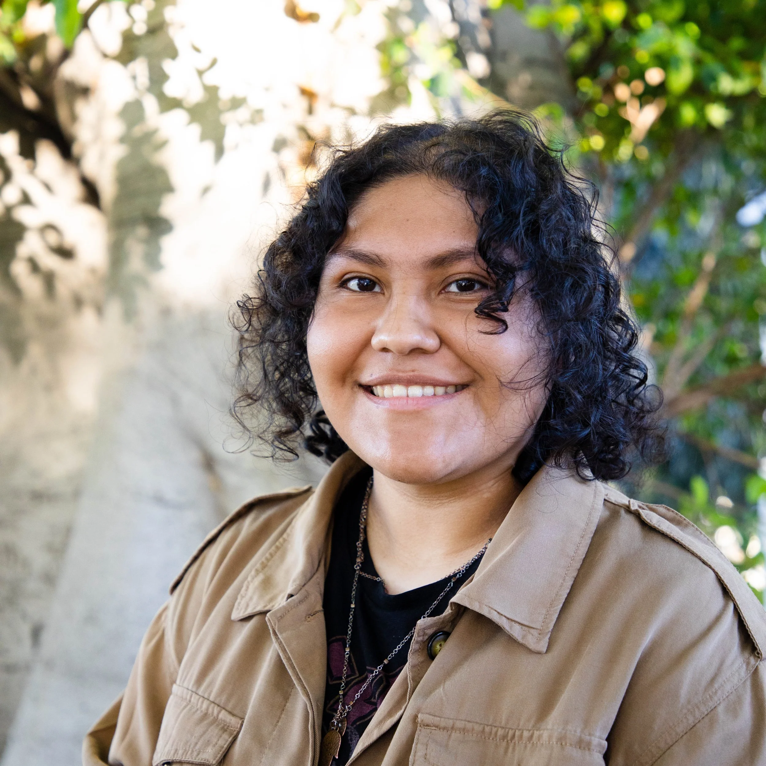 Portrait of a smiling woman with short curly black hair, wearing a tan jacket, standing outdoors with trees and greenery in the background.