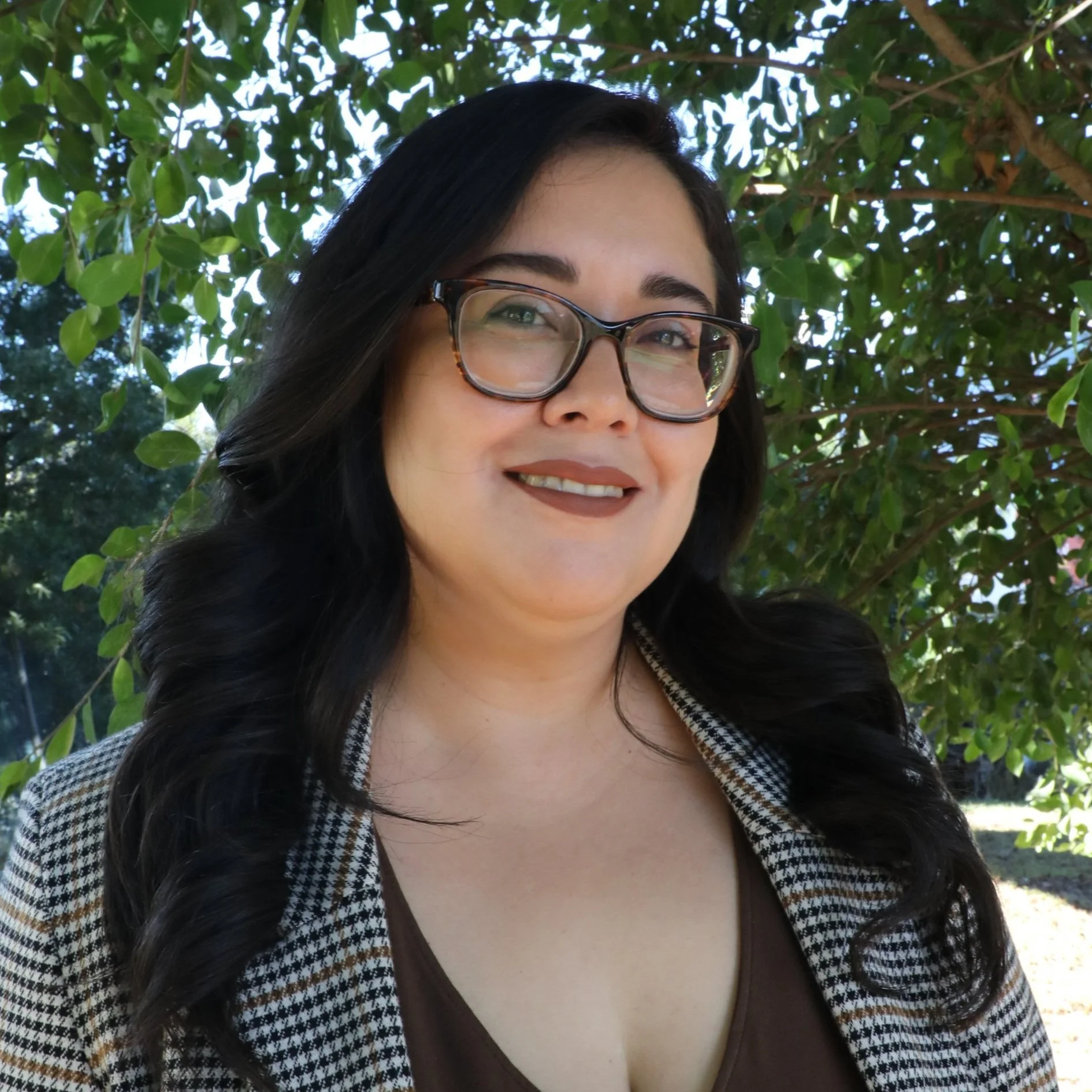 A woman with long black hair, wearing glasses and a houndstooth blazer, smiling outdoors in front of green leafy trees.