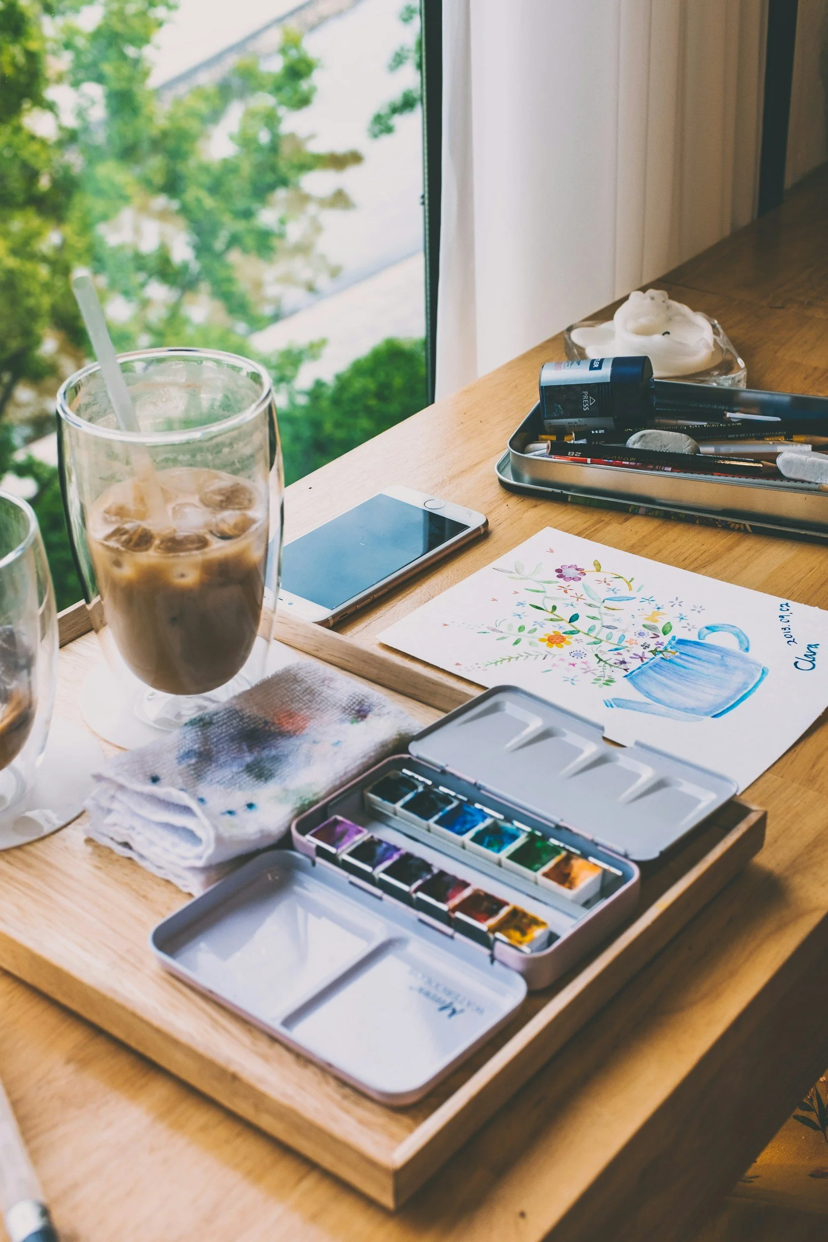 A wooden table near a window with an iced coffee, watercolor paints, a watercolor painting, a smartphone, and art supplies.